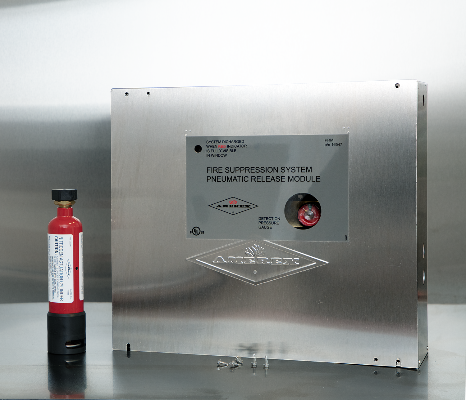 A stainless steel fire suppression system control box labeled “Pneumatic Release Module” sits next to a small red fire suppressant canister on a metallic surface.