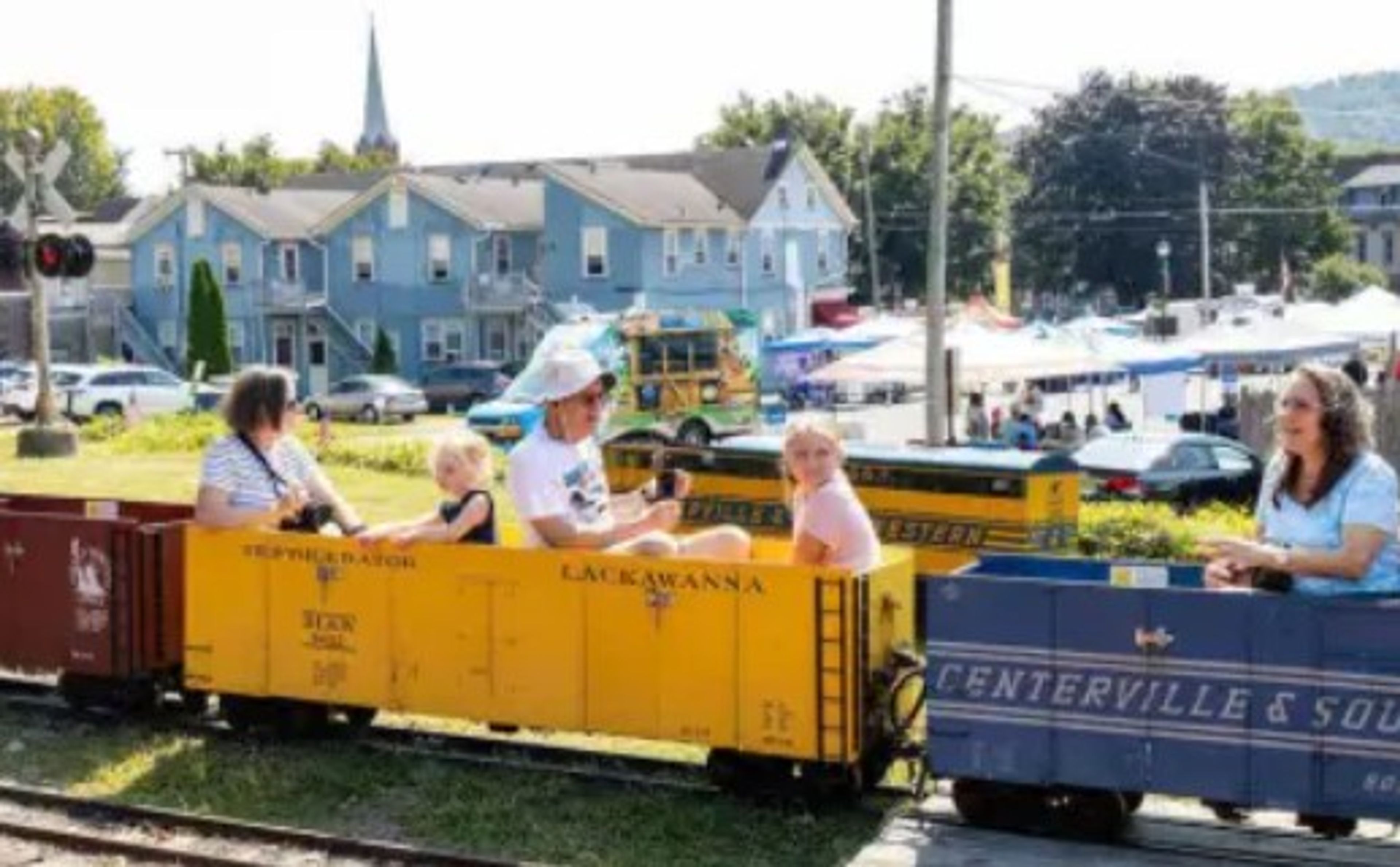 Adults and children ride a small, colorful train at an outdoor fair on a sunny day, with blue buildings, tents, and festival-goers in the background.