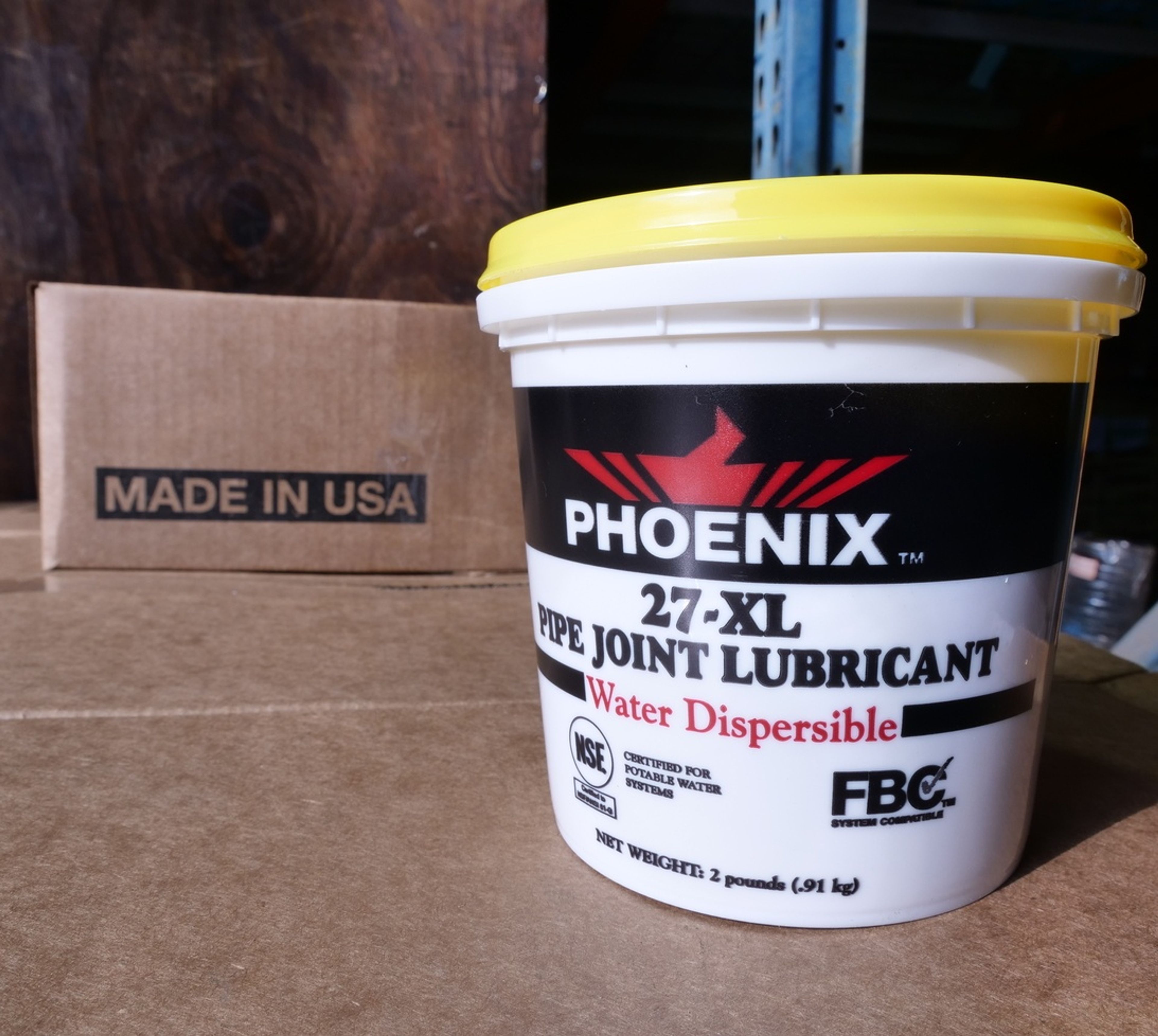 A white tub of Phoenix 27-XL Pipe Joint Lubricant with a yellow lid sits on a cardboard box. Another box labeled “MADE IN USA” is in the background.