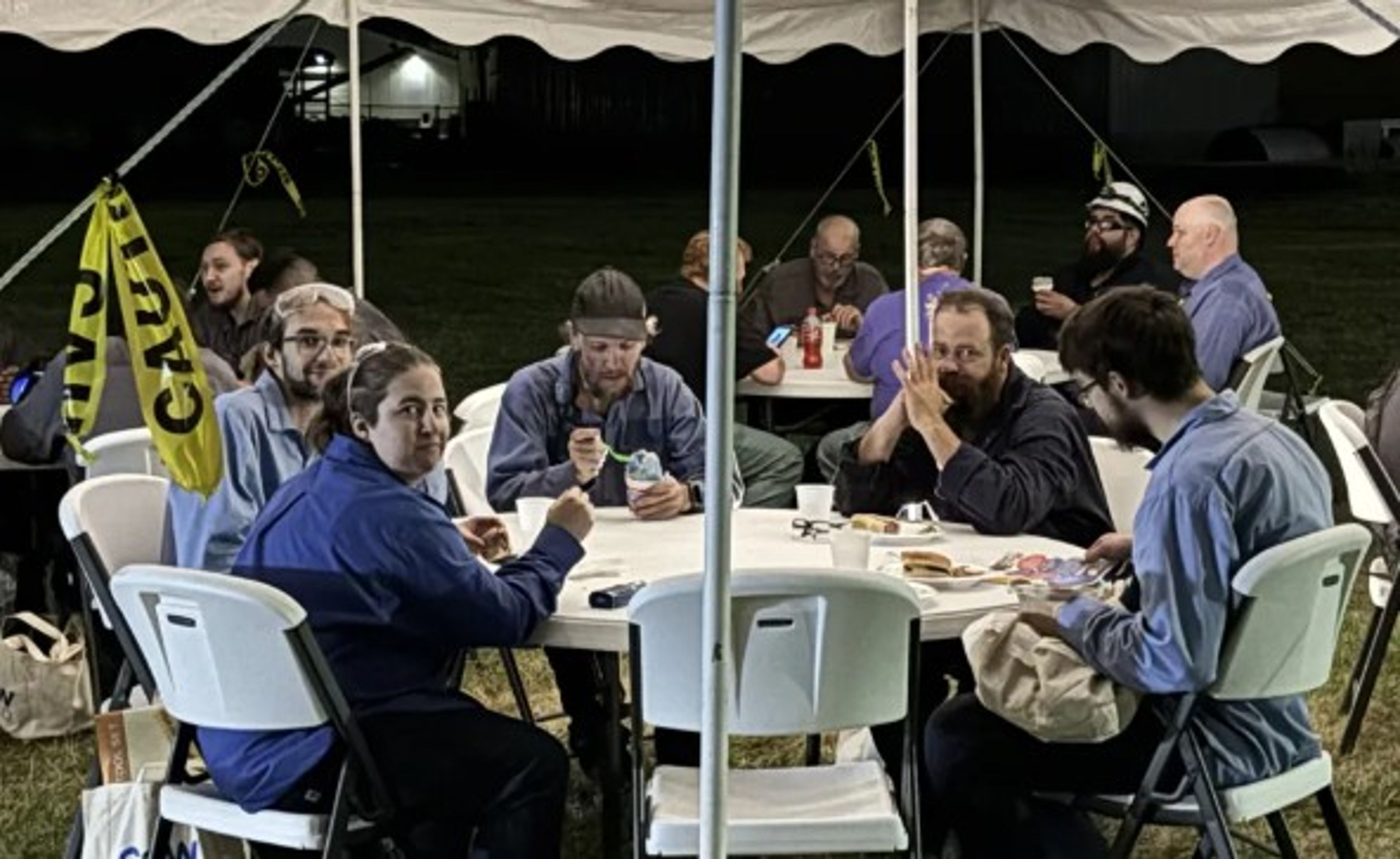 A group of people sitting at round tables under a tent at night, eating and talking. Some are wearing blue shirts, and a yellow “Caution” flag is visible. The scene appears to be an outdoor gathering or event.
