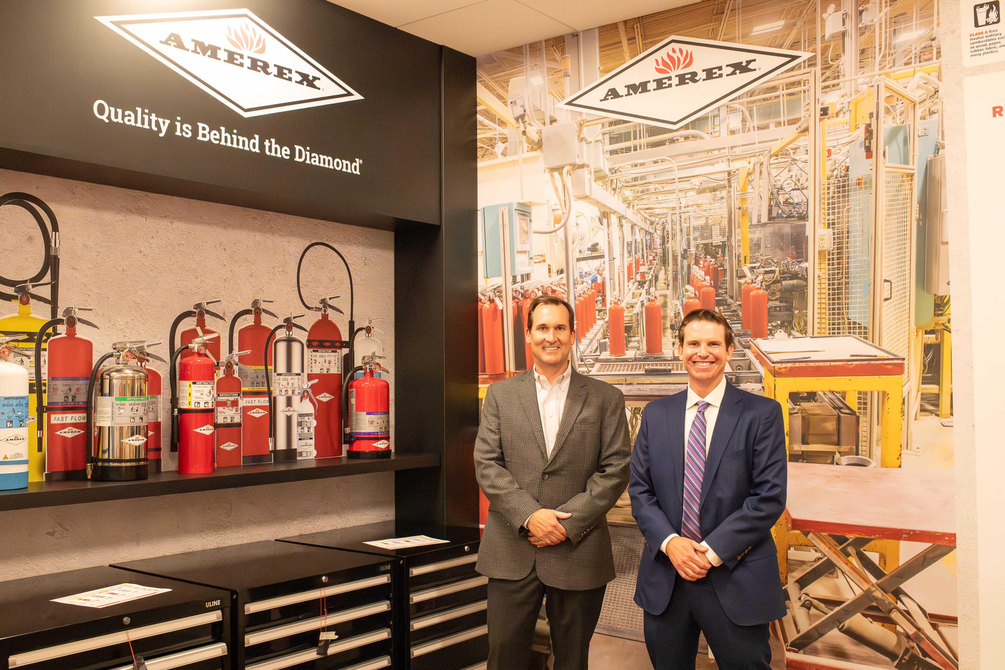 Two men in business suits stand smiling in front of an Amerex display with various fire extinguishers and a large factory photo. A sign reads, “Quality is Behind the Diamond.”.