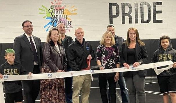A group of people, including children and adults, smile and hold a ribbon at an indoor ribbon-cutting ceremony. The words “Joseph Firth Youth Center” and “PRIDE” are visible on the wall behind them.