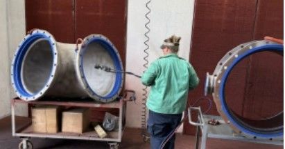 A person in a green shirt uses a tool to clean or inspect the inside of a large metal pipe in an industrial setting, with equipment and boxes nearby.