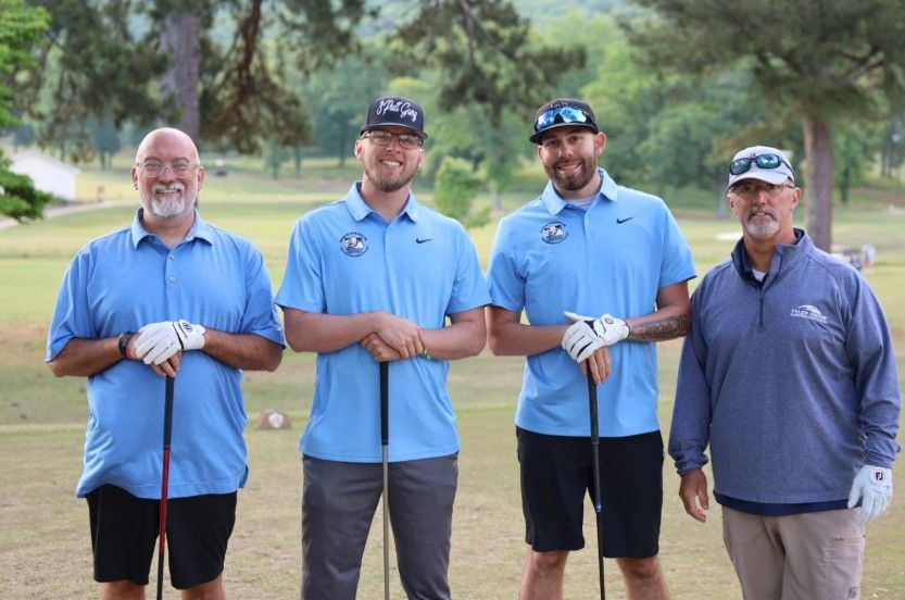 Four men stand on a golf course, smiling at the camera. Three wear matching light blue shirts and golf gloves, while the fourth wears a blue pullover and cap. They each hold golf clubs. Trees and green grass are in the background.