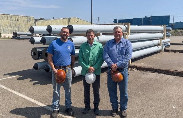 Three men wearing work shirts and holding hard hats stand in front of stacked utility poles in an outdoor industrial area on a sunny day.