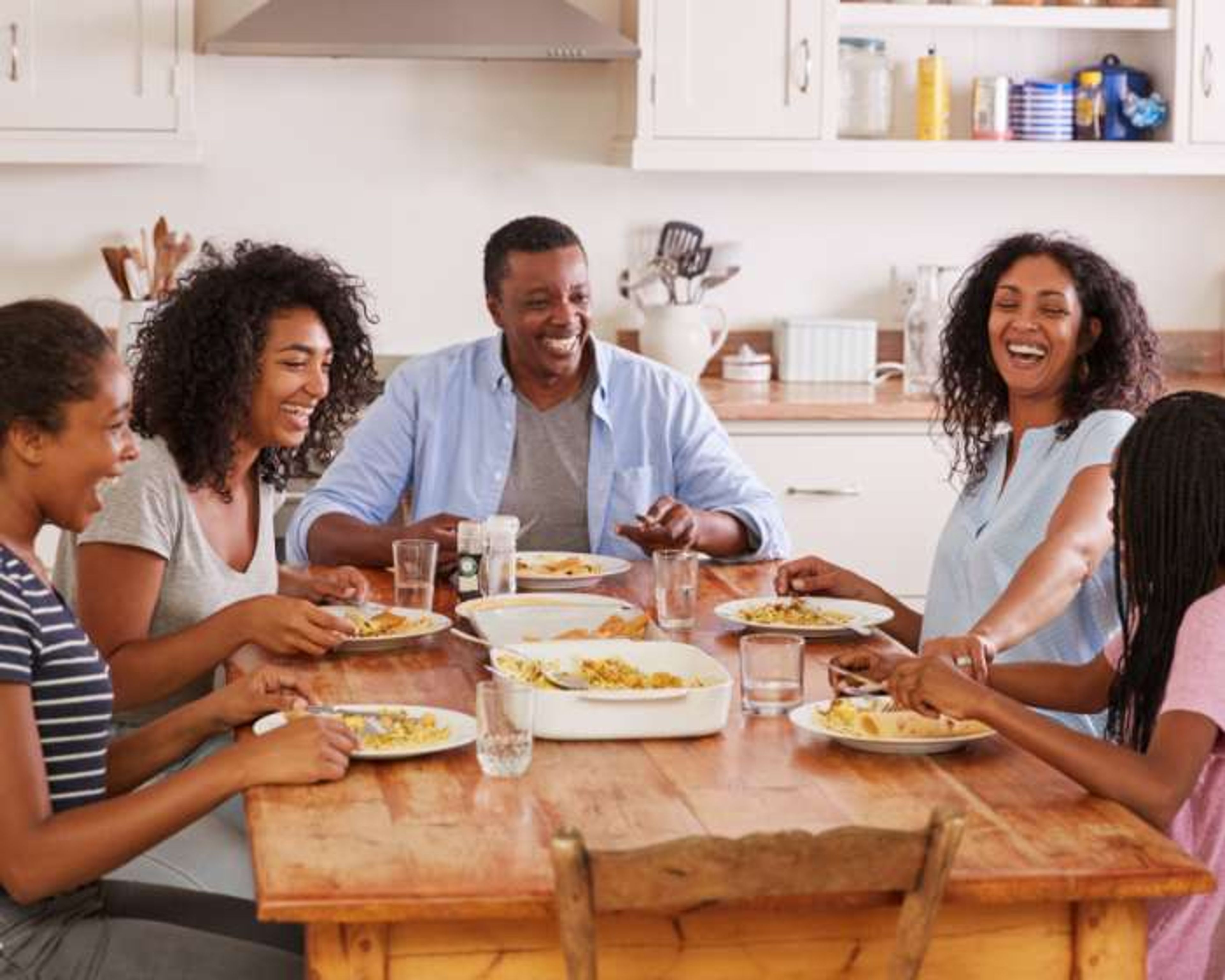 A family of five sits around a wooden table, smiling and laughing while eating a meal together in a bright kitchen. Plates and glasses are on the table.