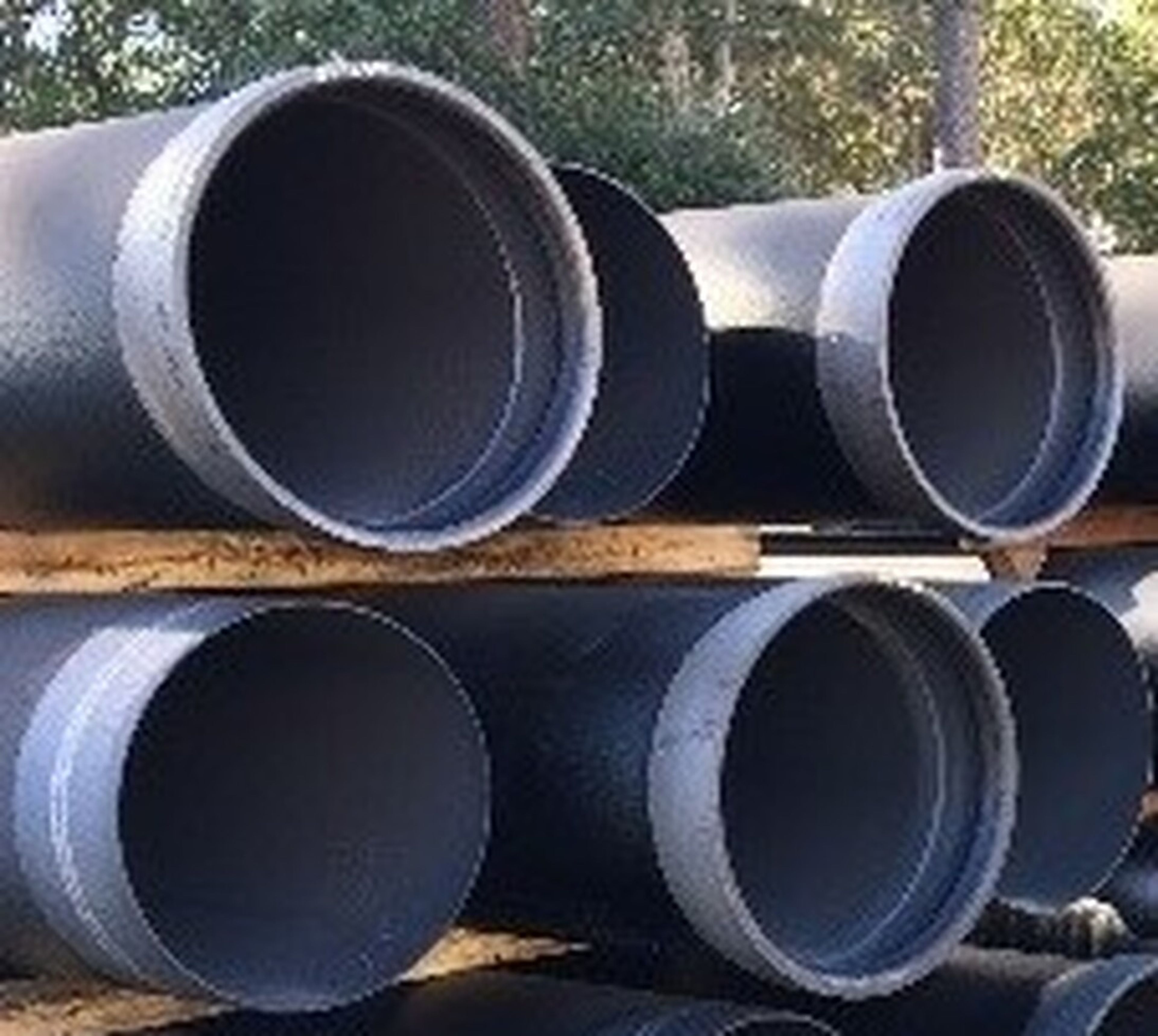 Stacks of large, black metal pipes are arranged horizontally outdoors with trees visible in the background.