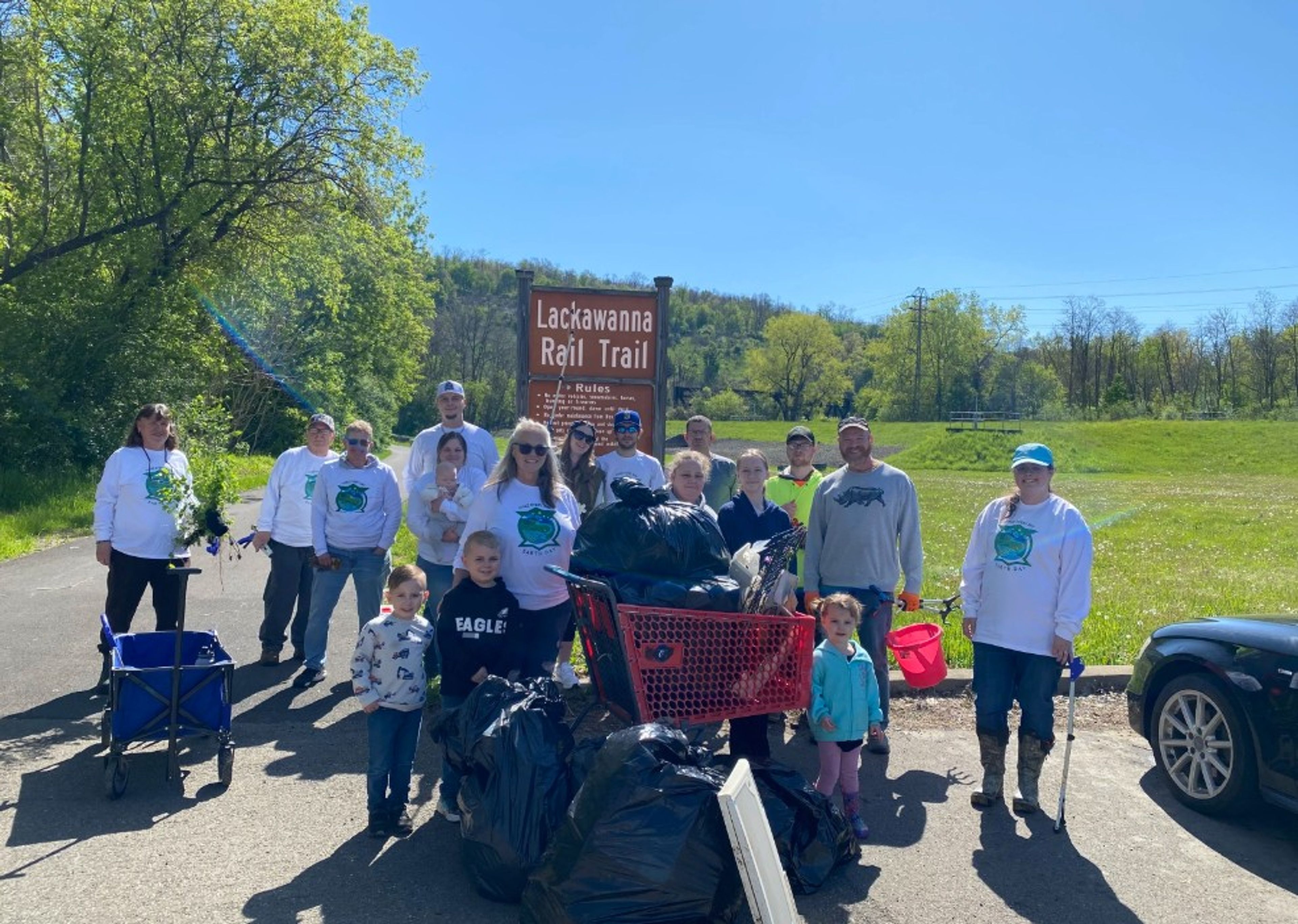 A group of people of various ages pose with collected trash bags in front of a “Lackawanna Rail Trail” sign on a sunny day, showing participation in a community cleanup event.