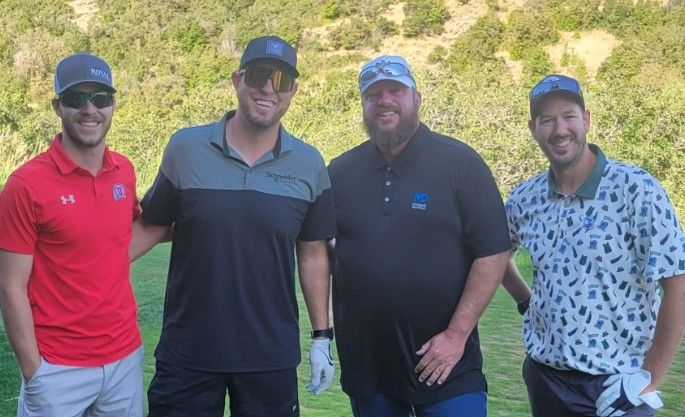 Four men stand together outdoors on a golf course, smiling at the camera. They are dressed in golf attire, wearing hats and sunglasses, with a green, wooded hillside in the background.