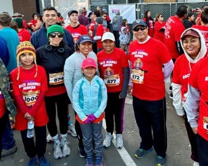 A group of people wearing red “Race Against Hunger” shirts and race bibs pose together outdoors at a race event. Some are smiling, and a young girl in blue stands in the front center.
