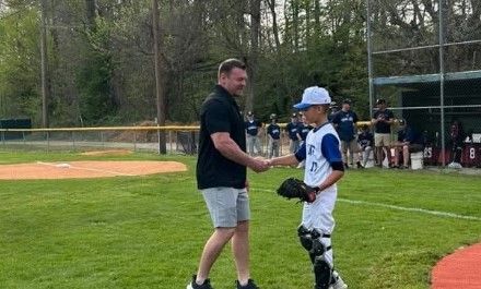 A man in a black shirt shakes hands with a young baseball player in catcher’s gear on a baseball field, with other players and spectators watching in the background.