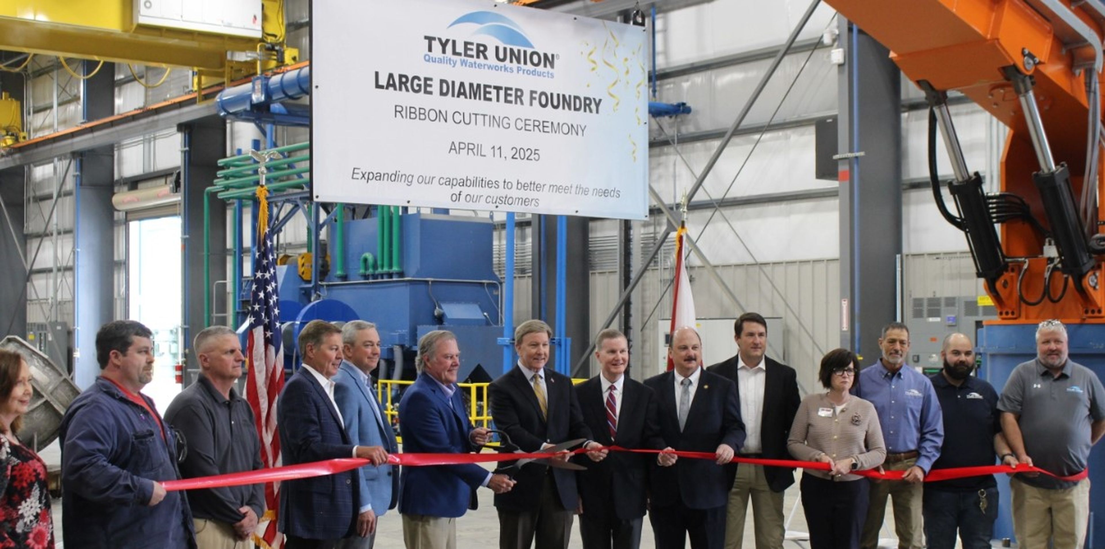 A group of people in business attire and uniforms cut a red ribbon at a foundry’s grand opening. A sign above reads “Tyler Union Large Diameter Foundry Ribbon Cutting Ceremony April 11, 2025.” Industrial equipment is visible.