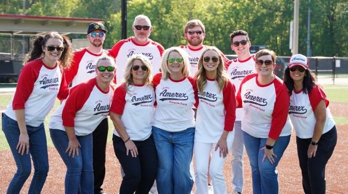 A group of twelve people wearing matching red and white “Amereex Financial Family” shirts pose and smile together on a sunny baseball field.