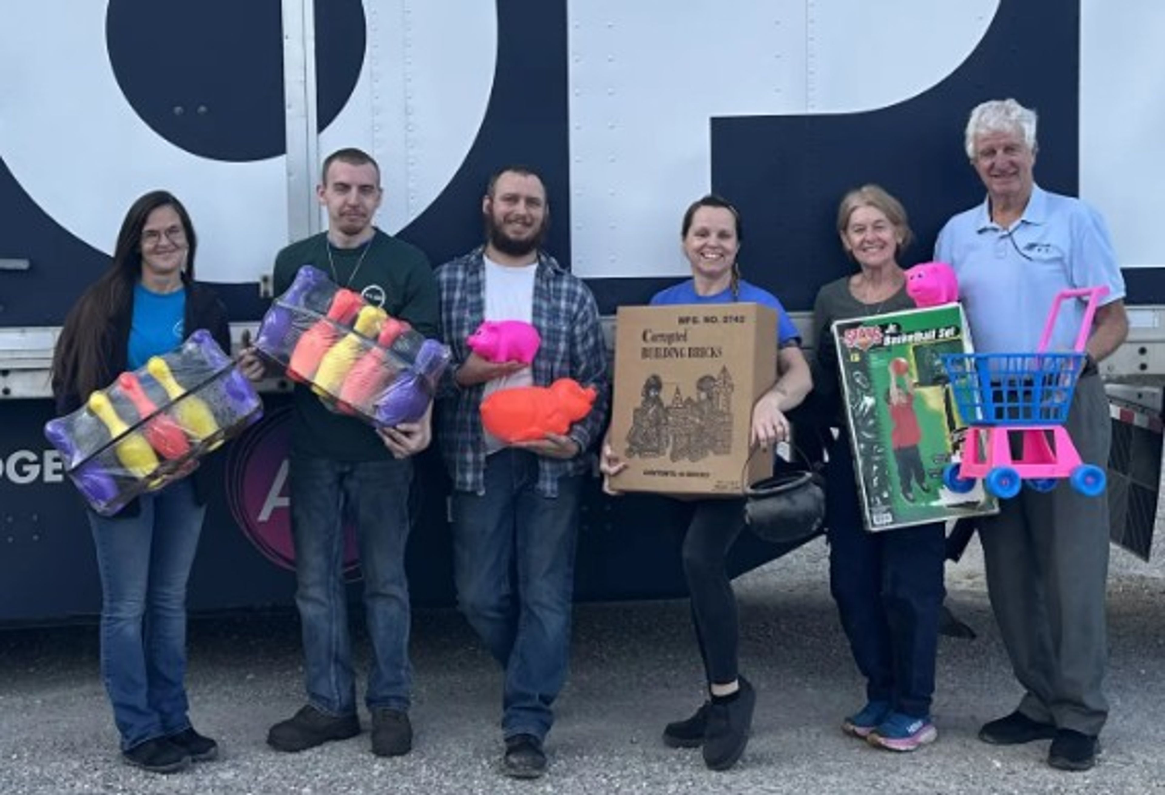 Six people stand smiling in front of a truck, each holding items such as yarn, a box, a toy stroller, and a toy kitchen set. It appears to be a volunteer or donation event.