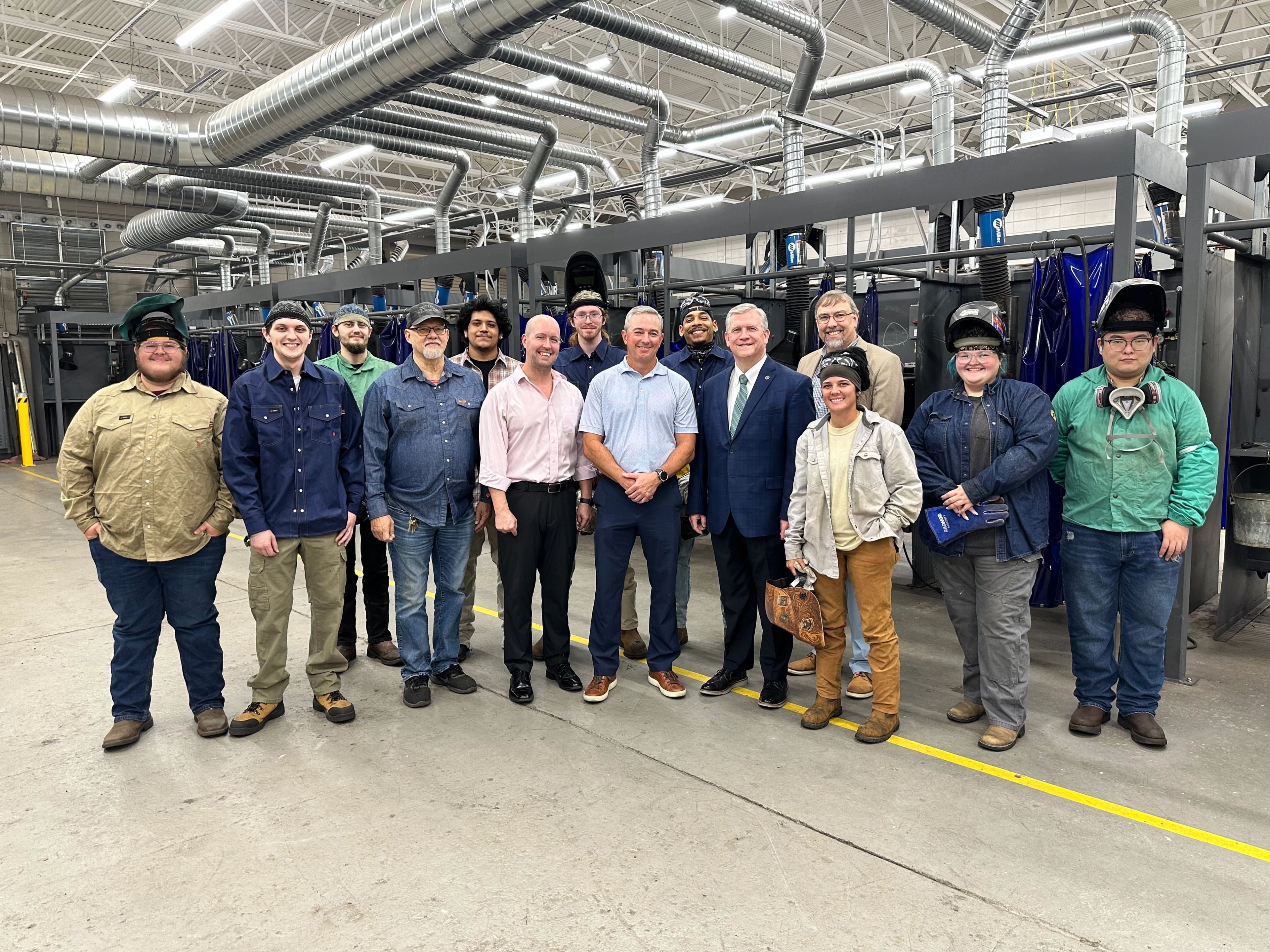 A group of people, some in business attire and others in work clothes and welding helmets, stand together inside a large industrial facility with machinery and ventilation ducts visible in the background.