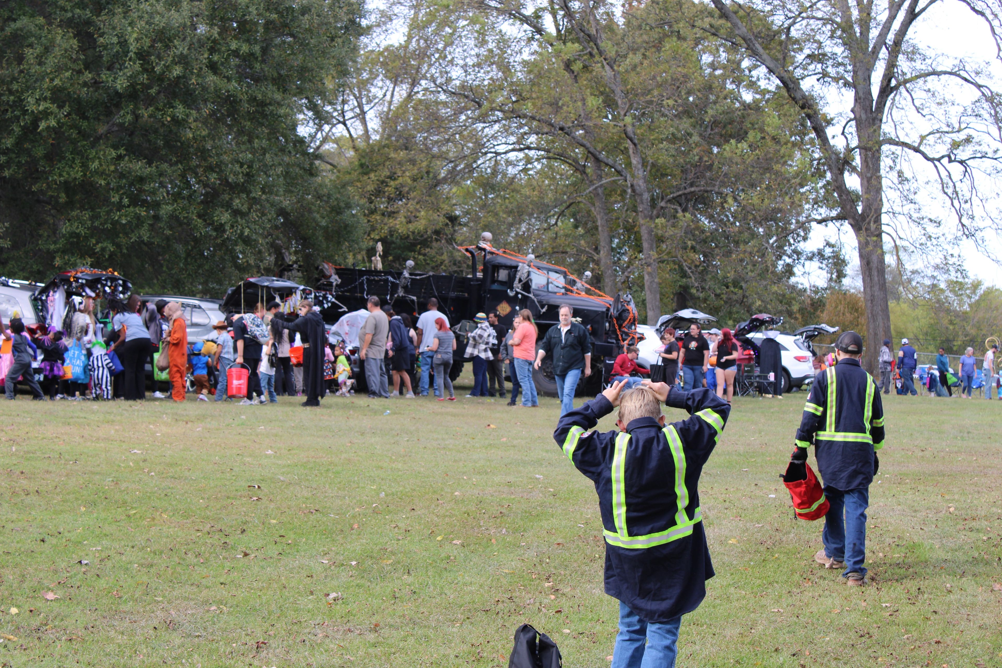 A crowd gathers at an outdoor event near parked vehicles. In the foreground, two children in safety vests hold bags, while others line up near trucks under tall trees on a grassy field.