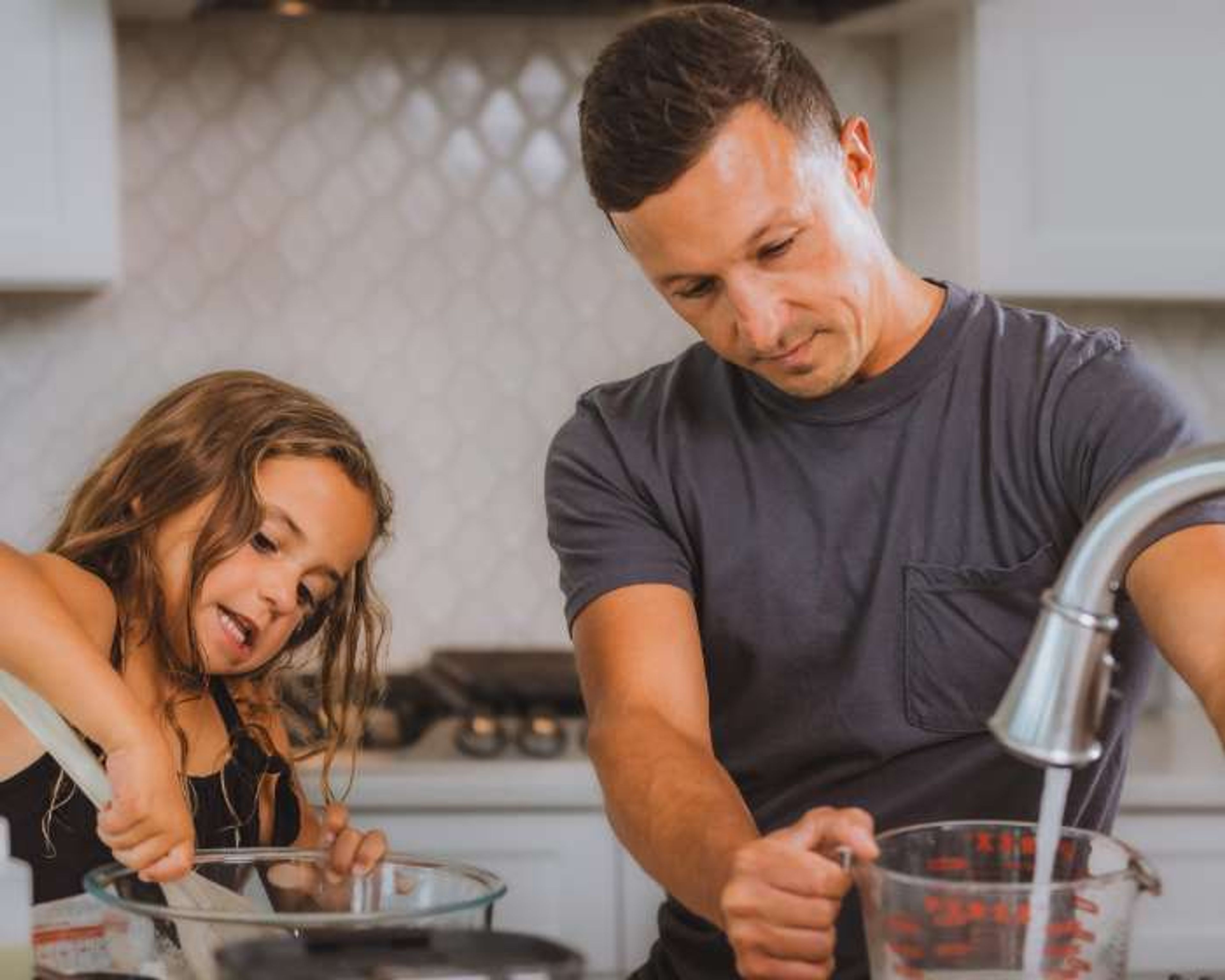 A man and a young girl cook together in a kitchen; the girl stirs a bowl while the man uses a measuring cup and fills it with water from the sink.
