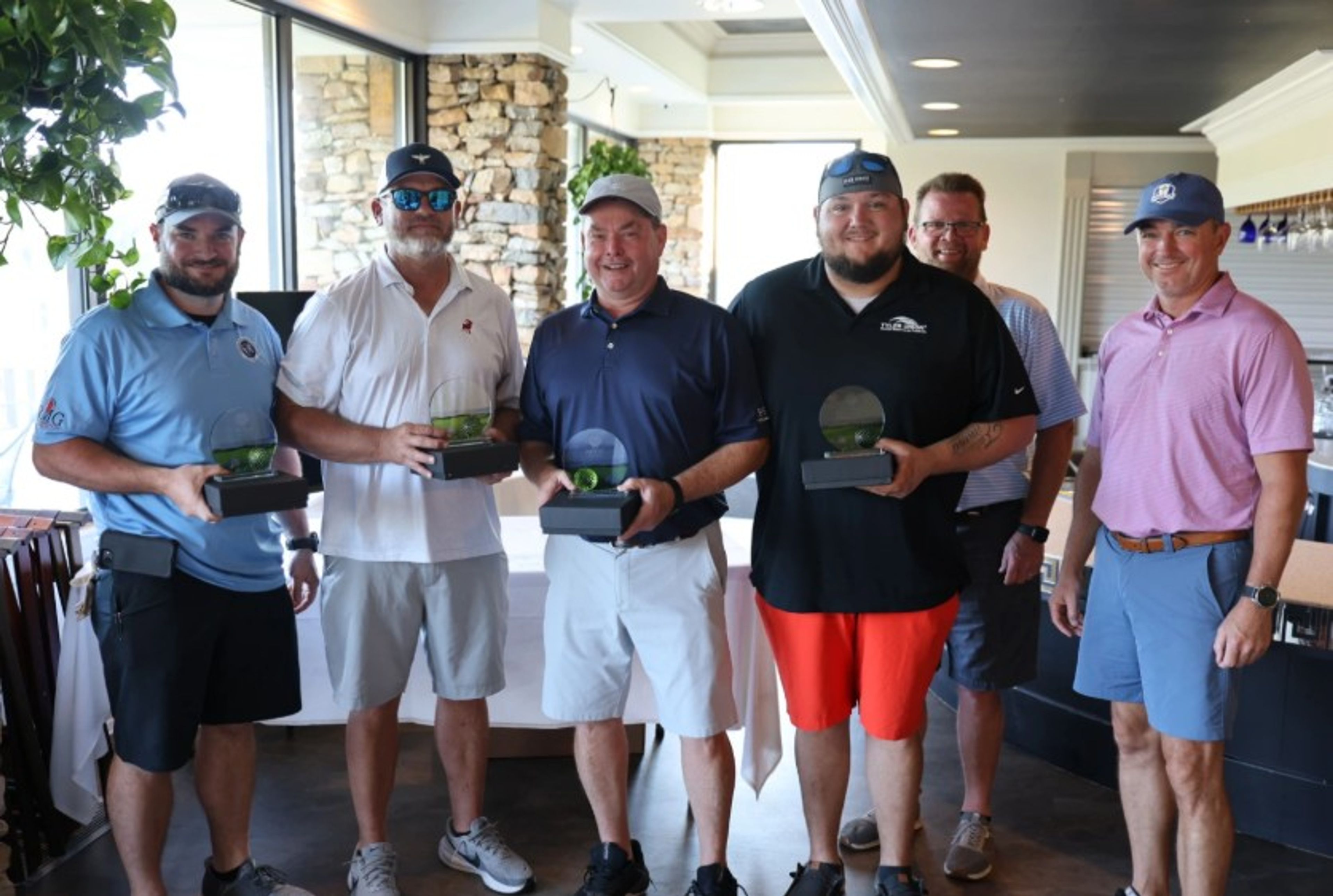 Six men pose indoors; four hold glass awards. They wear casual golf attire, including polos, shorts, and hats. The background shows a stone wall and large window with natural light coming in.