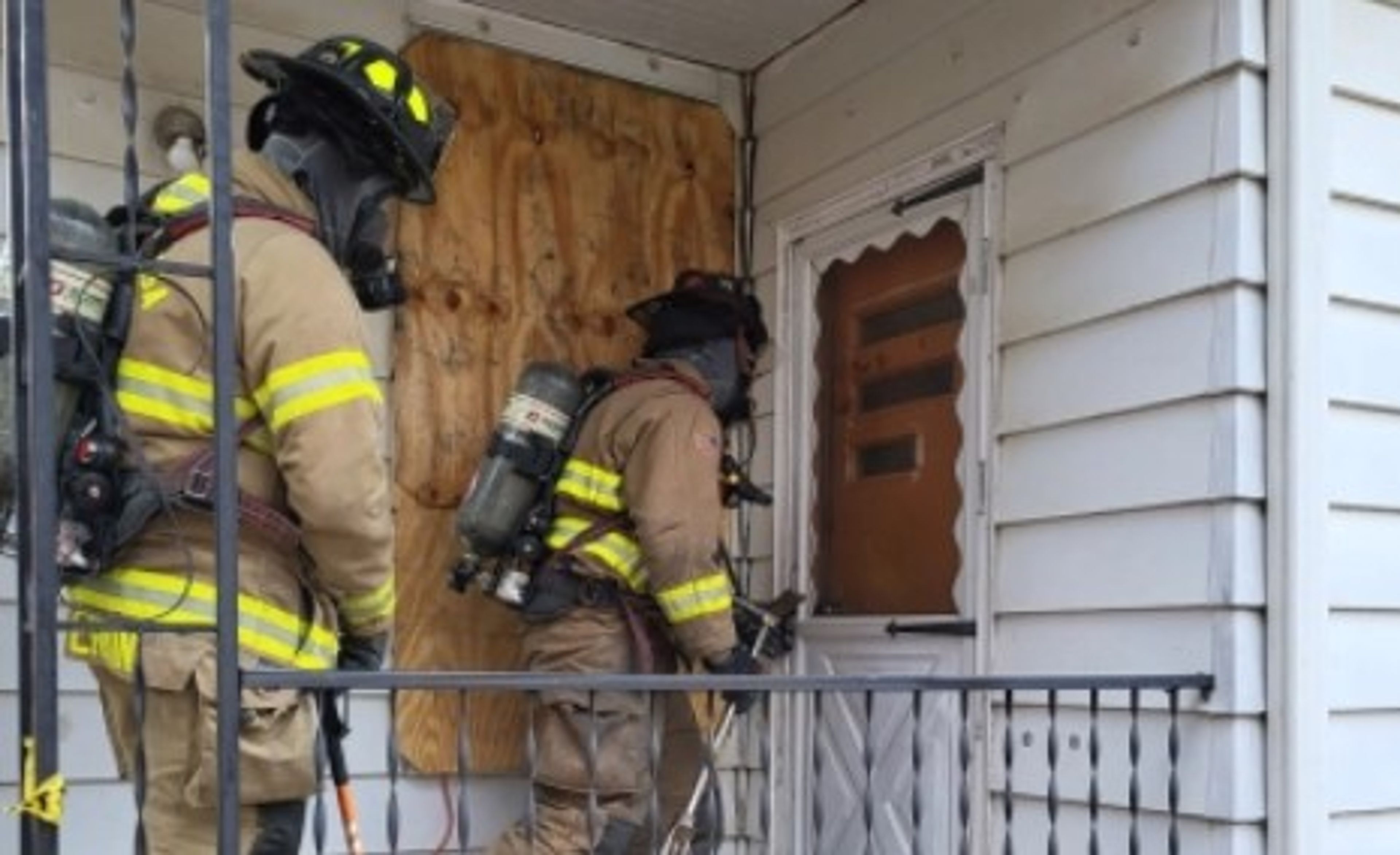 Two firefighters in full gear stand on a porch. One uses a tool to open a door, while the other stands behind. A window next to the door is boarded up with plywood.