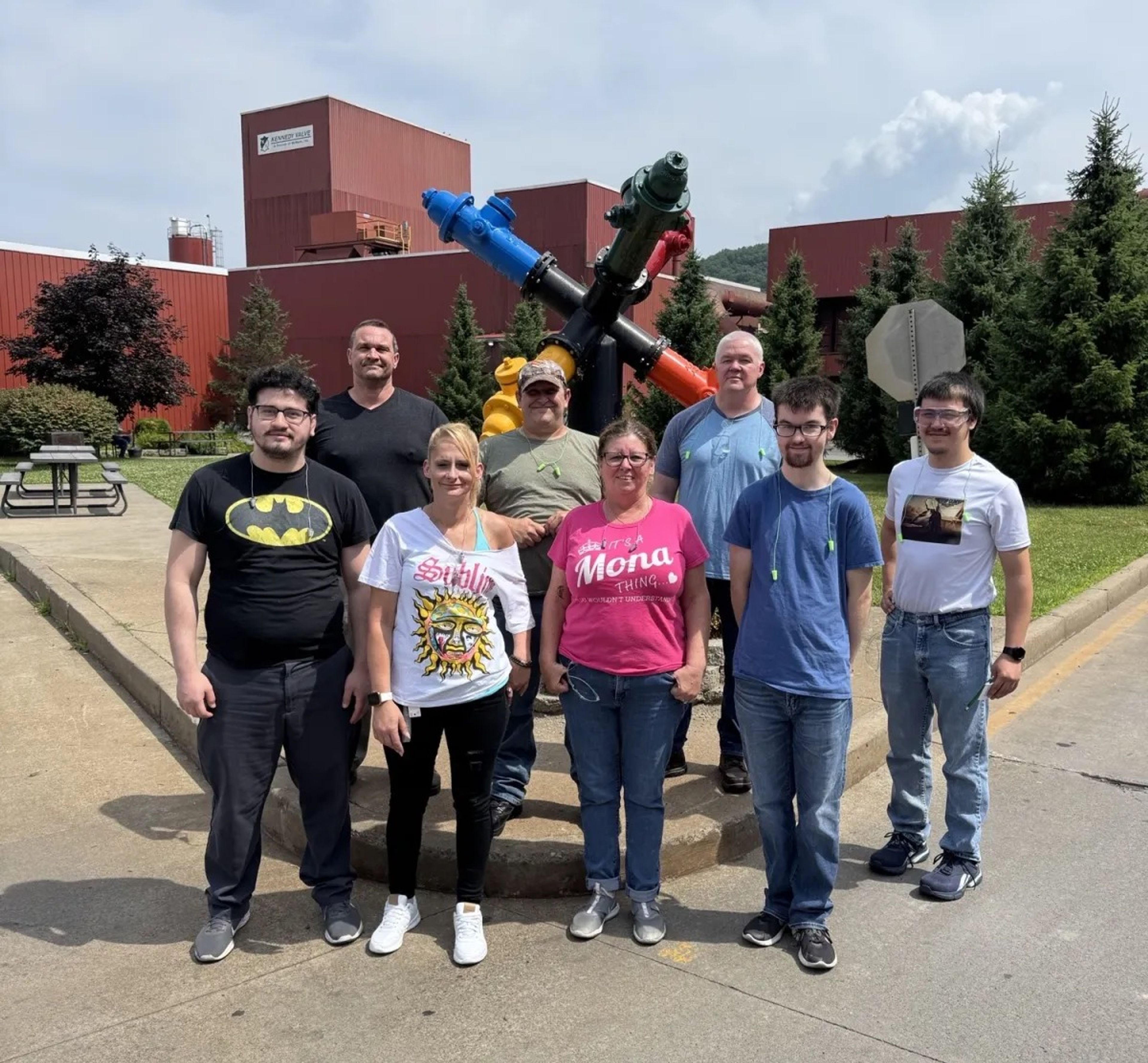 Eight people stand outdoors in front of a colorful hydrant sculpture, with red buildings and trees in the background. The group poses and smiles on a sunny day.