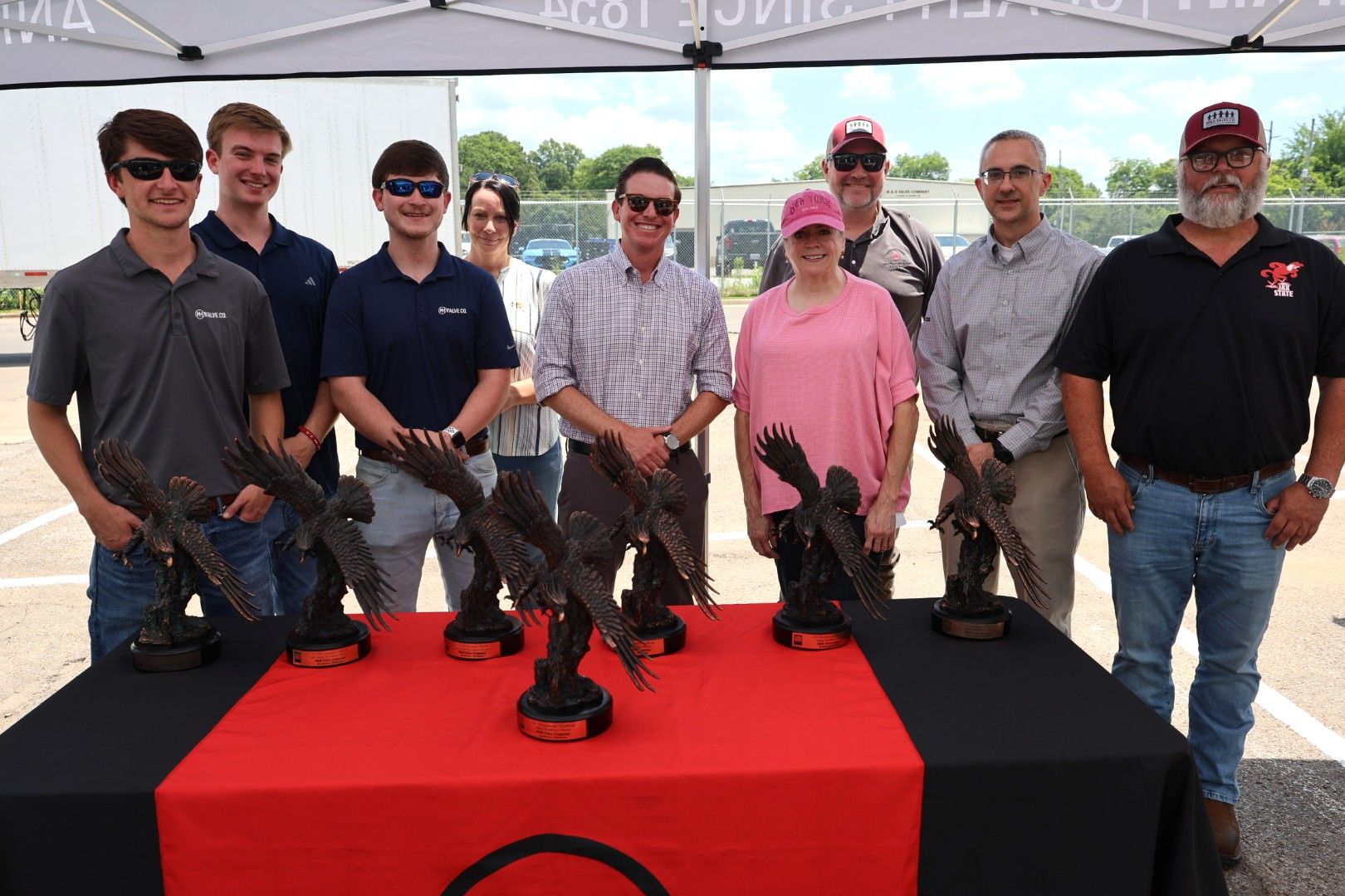 Nine people stand behind a table with five bird trophies on it, under a canopy outdoors. Most are smiling and wearing casual clothes, sunglasses, and hats. The table is covered with a red and black cloth.
