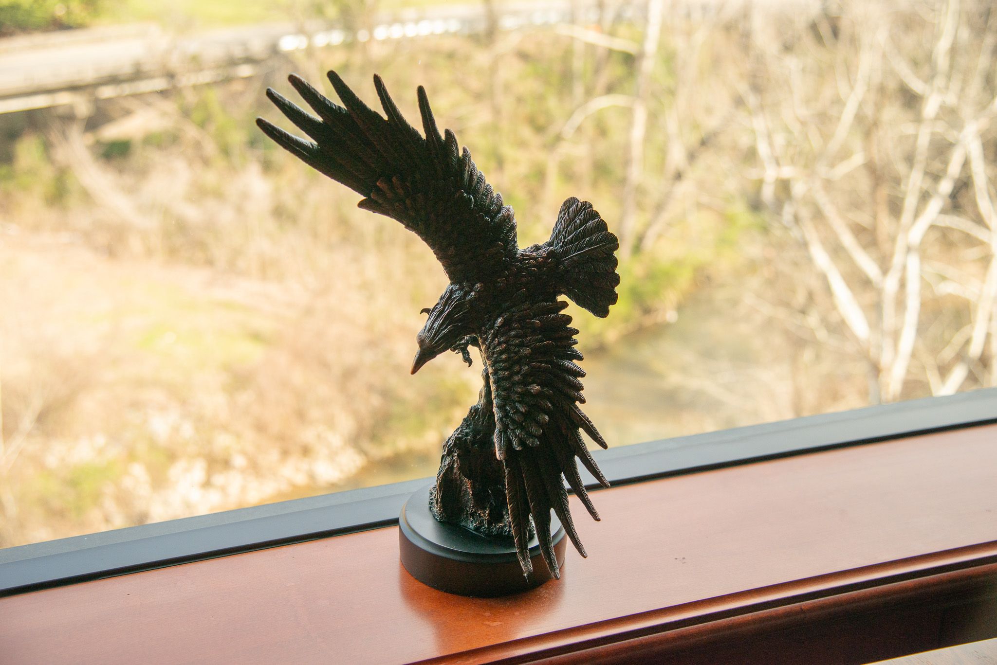 A black raven sculpture with wings spread sits on a wooden windowsill. A blurred outdoor scene with trees and a river is visible through the window in the background.
