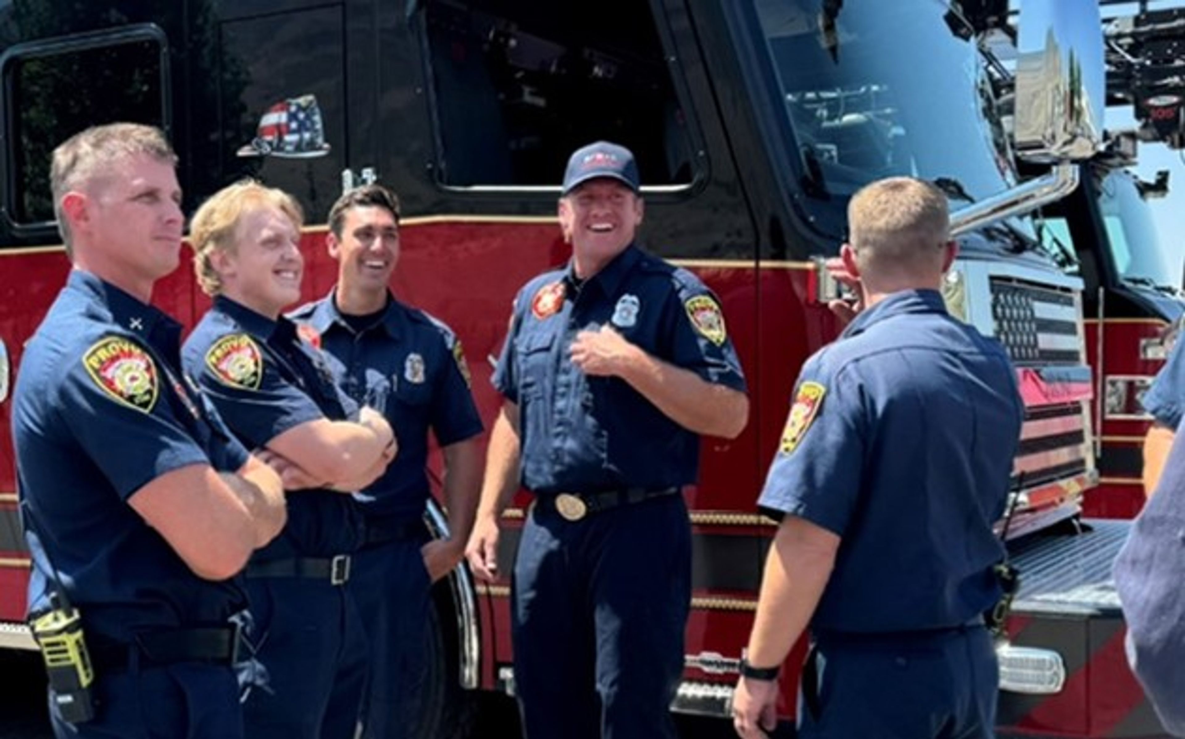 Five firefighters in uniform stand and chat, smiling, in front of a red fire truck on a sunny day.