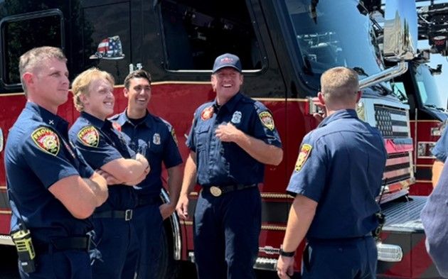 Five firefighters in uniform stand and chat, smiling, in front of a red fire truck on a sunny day.