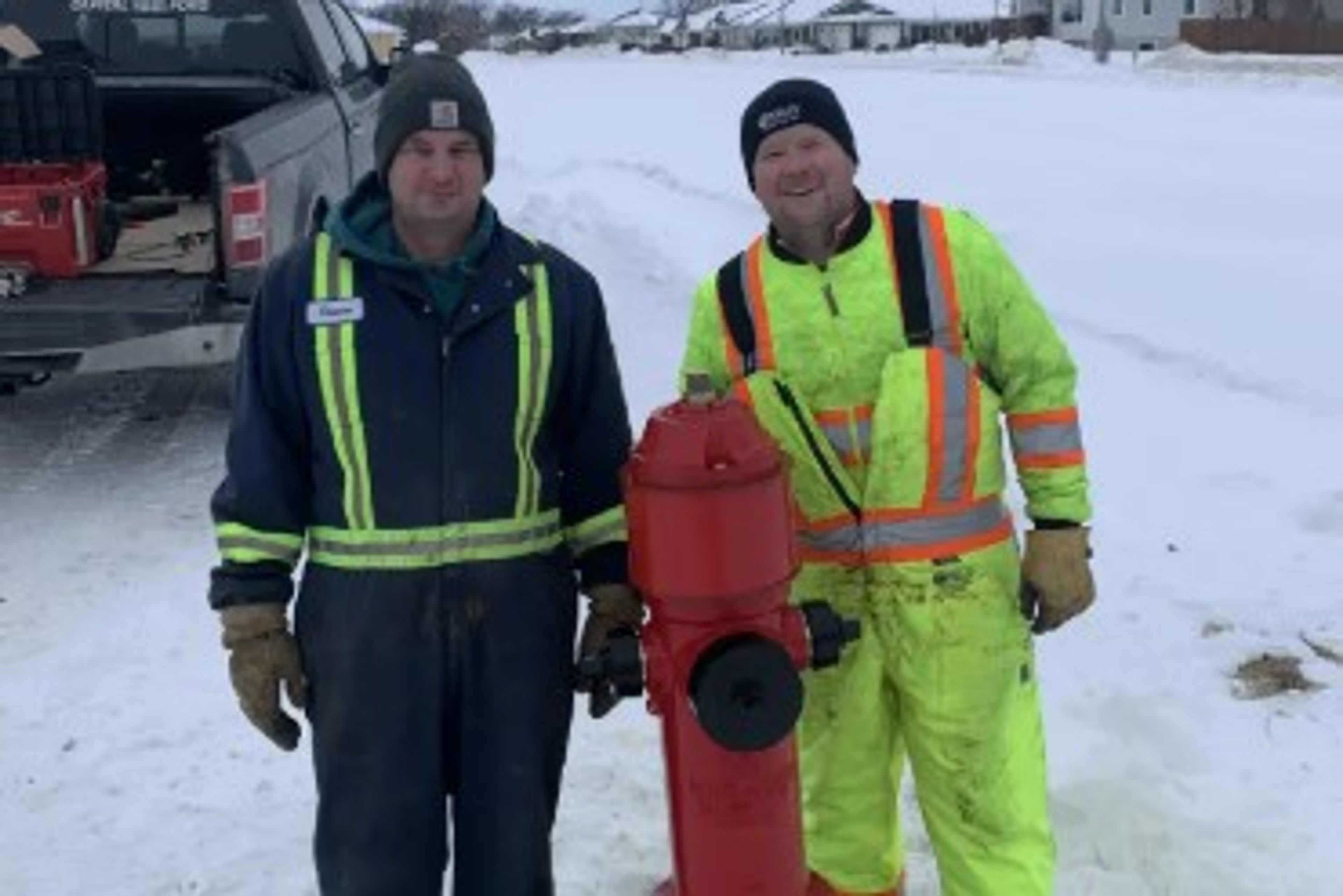 Two workers in winter gear stand by a red fire hydrant on a snowy street. A pickup truck with tools is parked behind them, and houses are visible in the background.