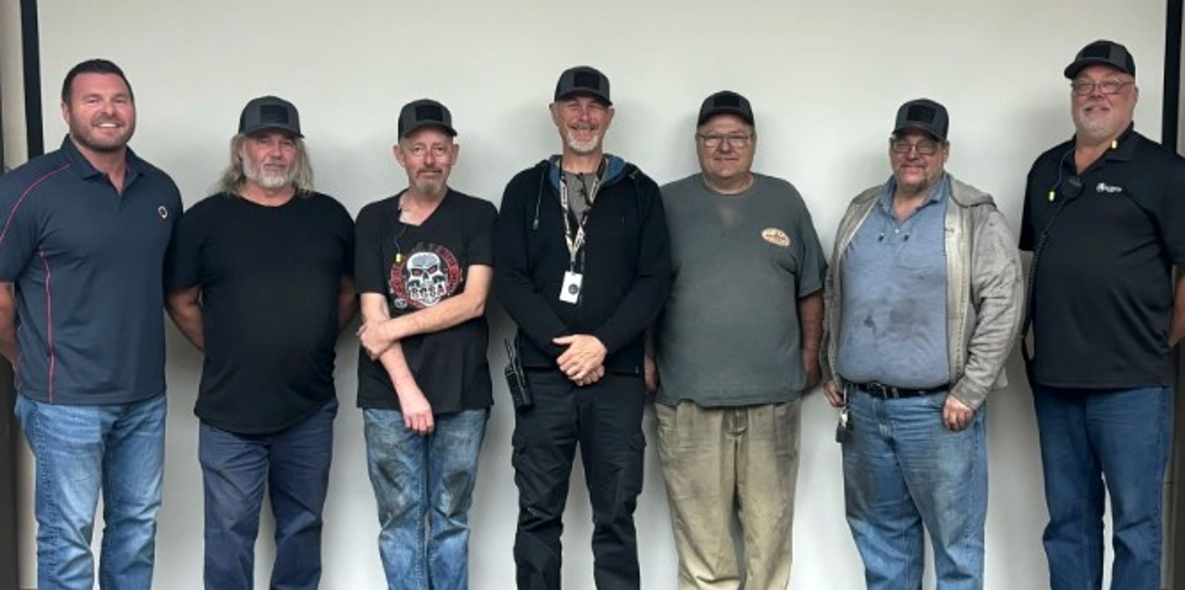 Seven men wearing hats stand in a row against a plain wall, smiling at the camera. Most are dressed casually in jeans and shirts, with two in collared shirts on each end.