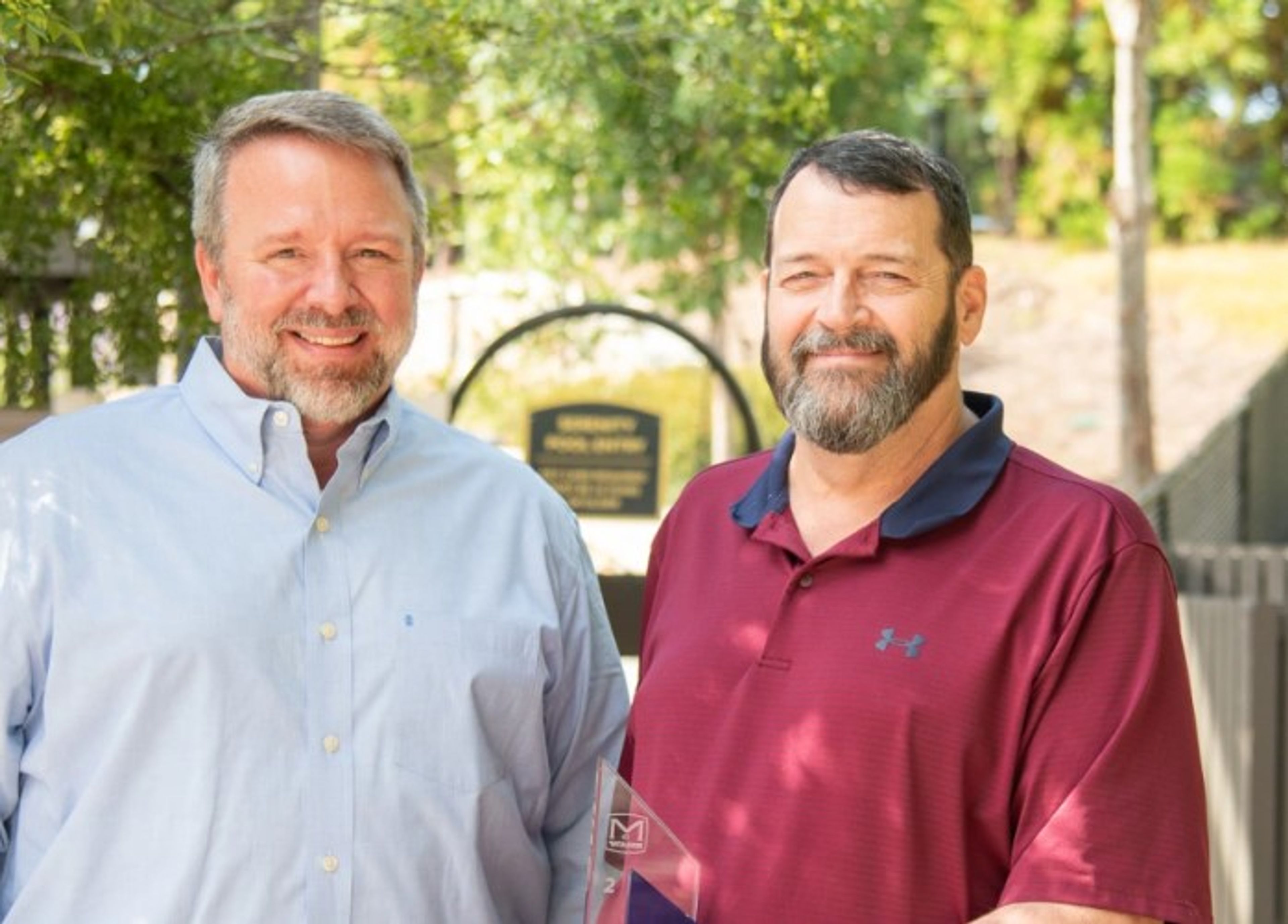 Two smiling men stand outdoors in front of a sign, with greenery and trees in the background. One man wears a light blue shirt, the other a maroon polo shirt.