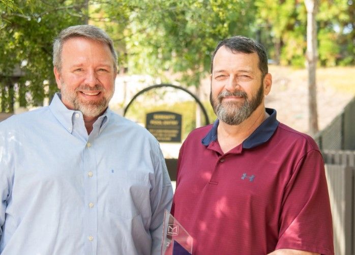 Two smiling men stand outdoors in front of a sign, with greenery and trees in the background. One man wears a light blue shirt, the other a maroon polo shirt.