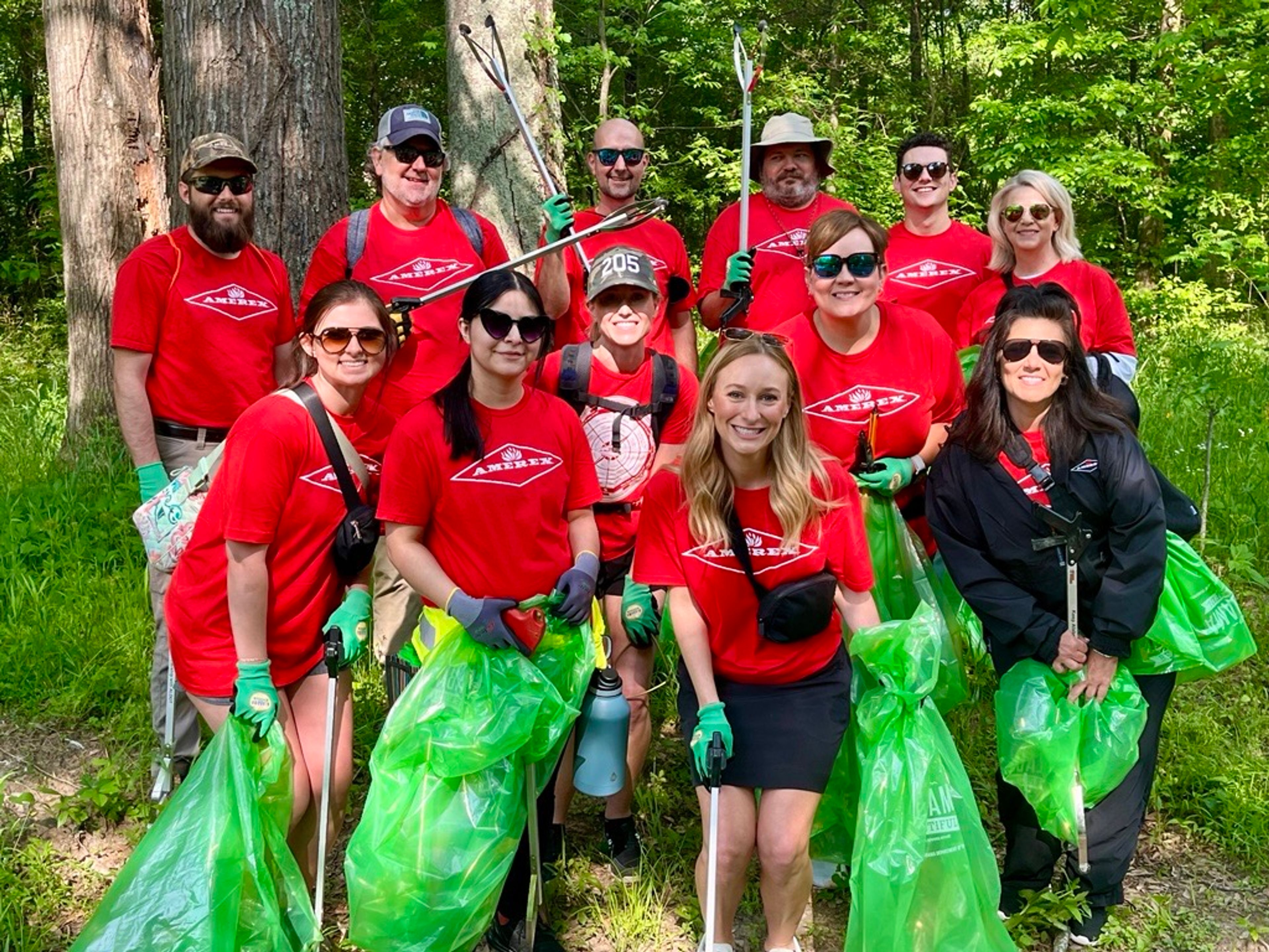 A group of people wearing red shirts and green gloves pose in a forest while holding green trash bags, appearing to participate in a community cleanup event.