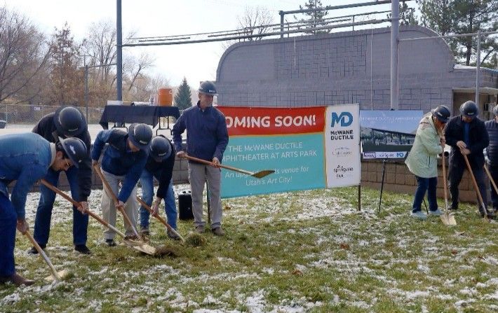 A group of people in hard hats and coats participate in a groundbreaking ceremony with shovels, standing on a patchy snowy lawn near a sign that says “COMING SOON!” for an amphitheater project.