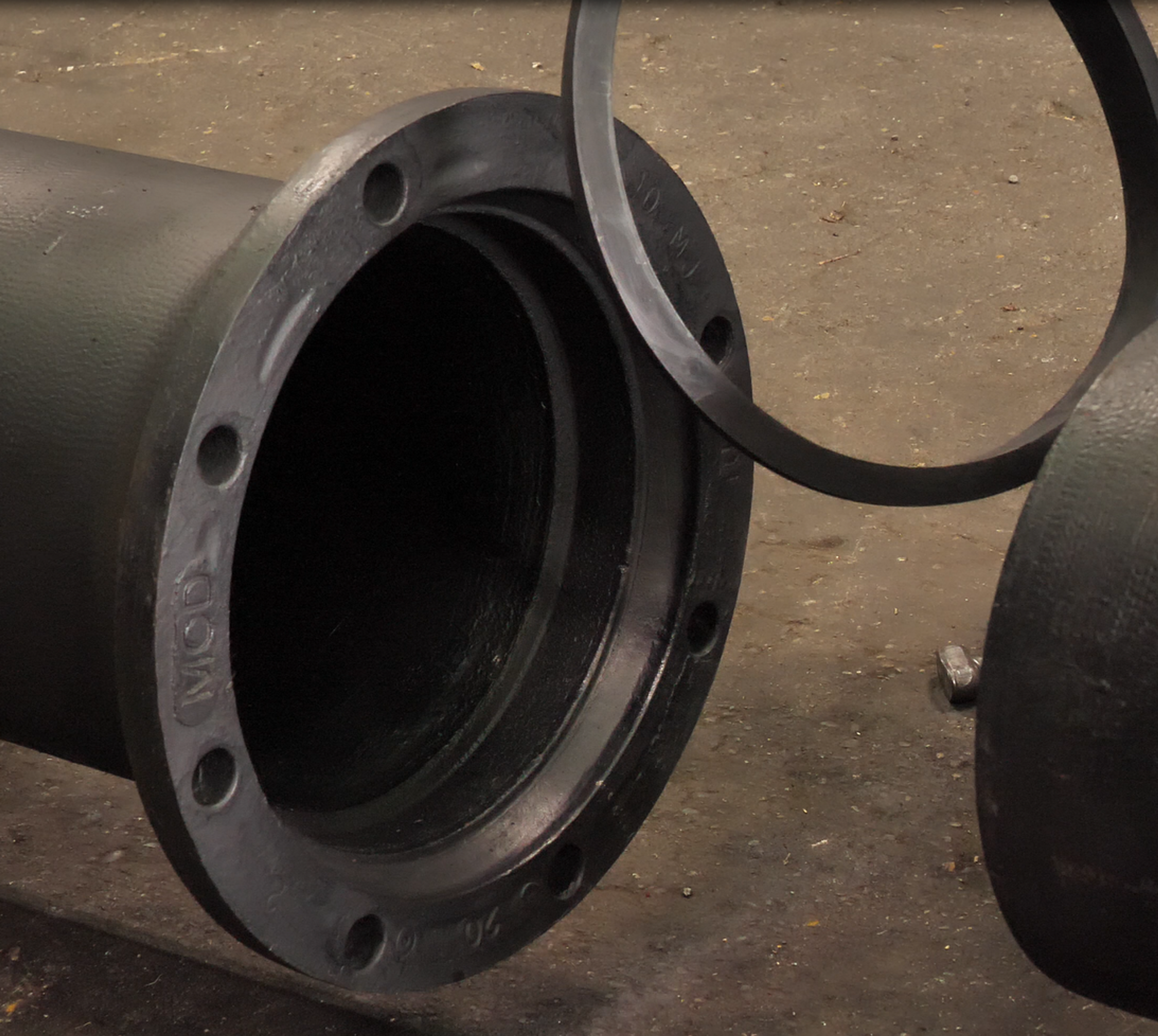 A close-up of two large, dark metal pipes with flanged ends on a workshop floor. One pipe is lying horizontally, while another pipe is positioned nearby.