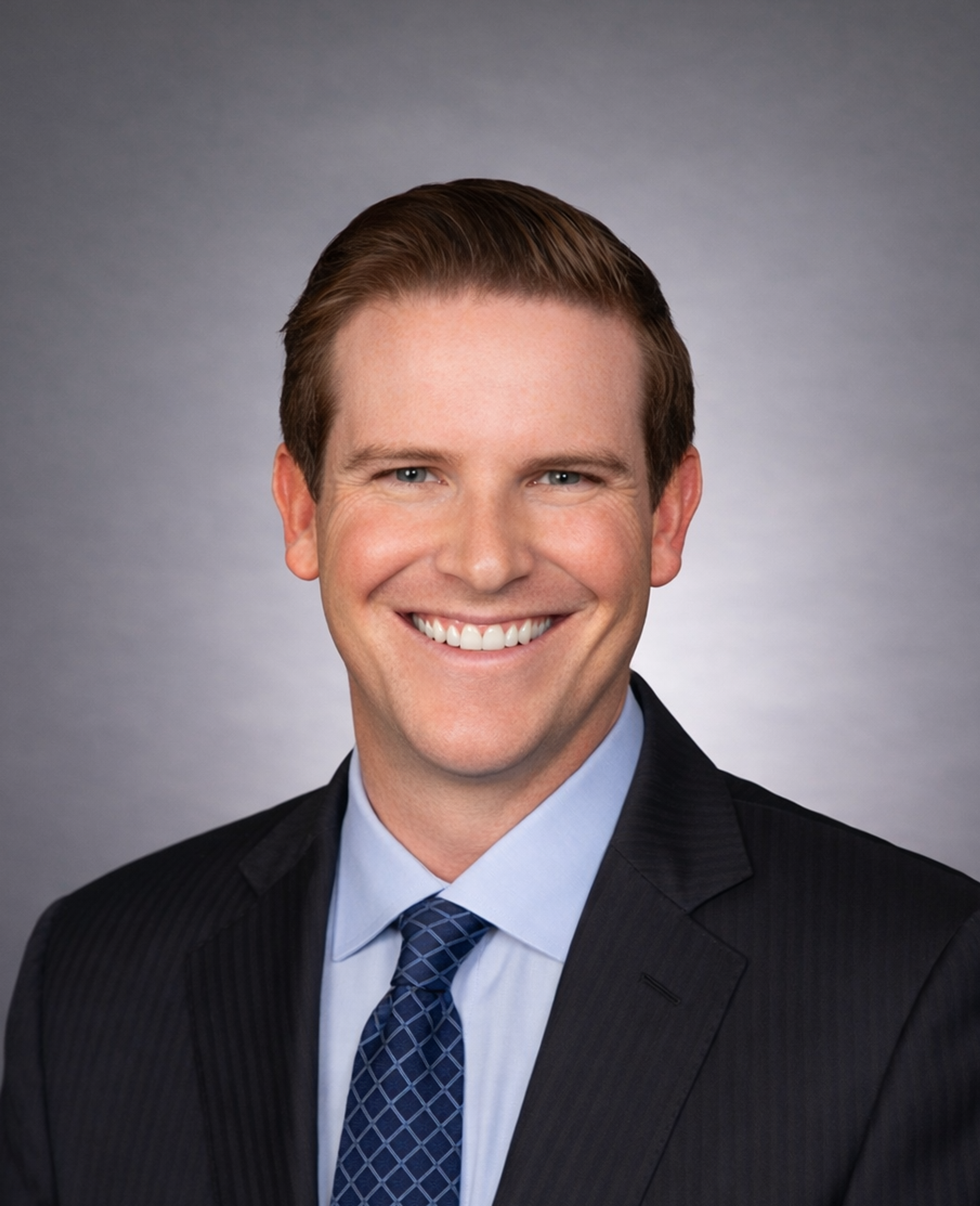 A man with short brown hair in a suit and blue tie smiles at the camera against a gray background.