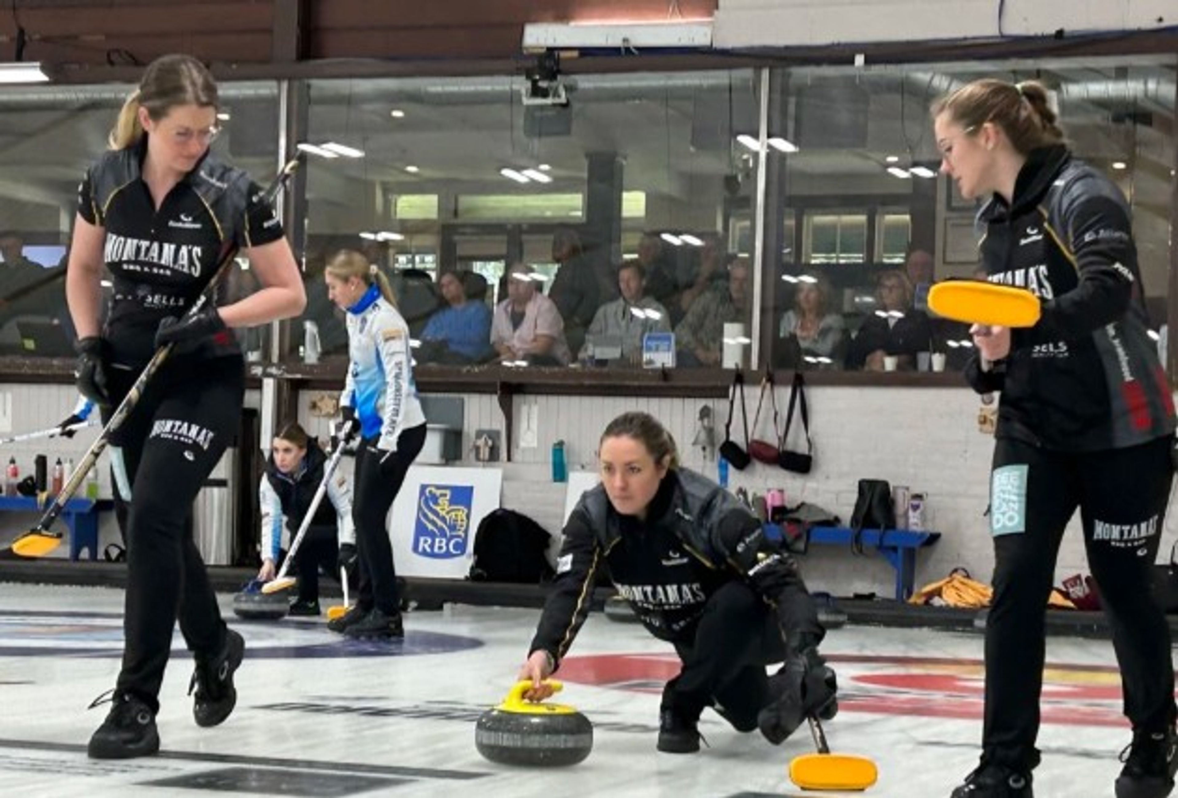Three women in black curling uniforms play on an indoor ice rink. One slides a stone while the others hold brooms, ready to sweep. Spectators watch through glass windows in the background.