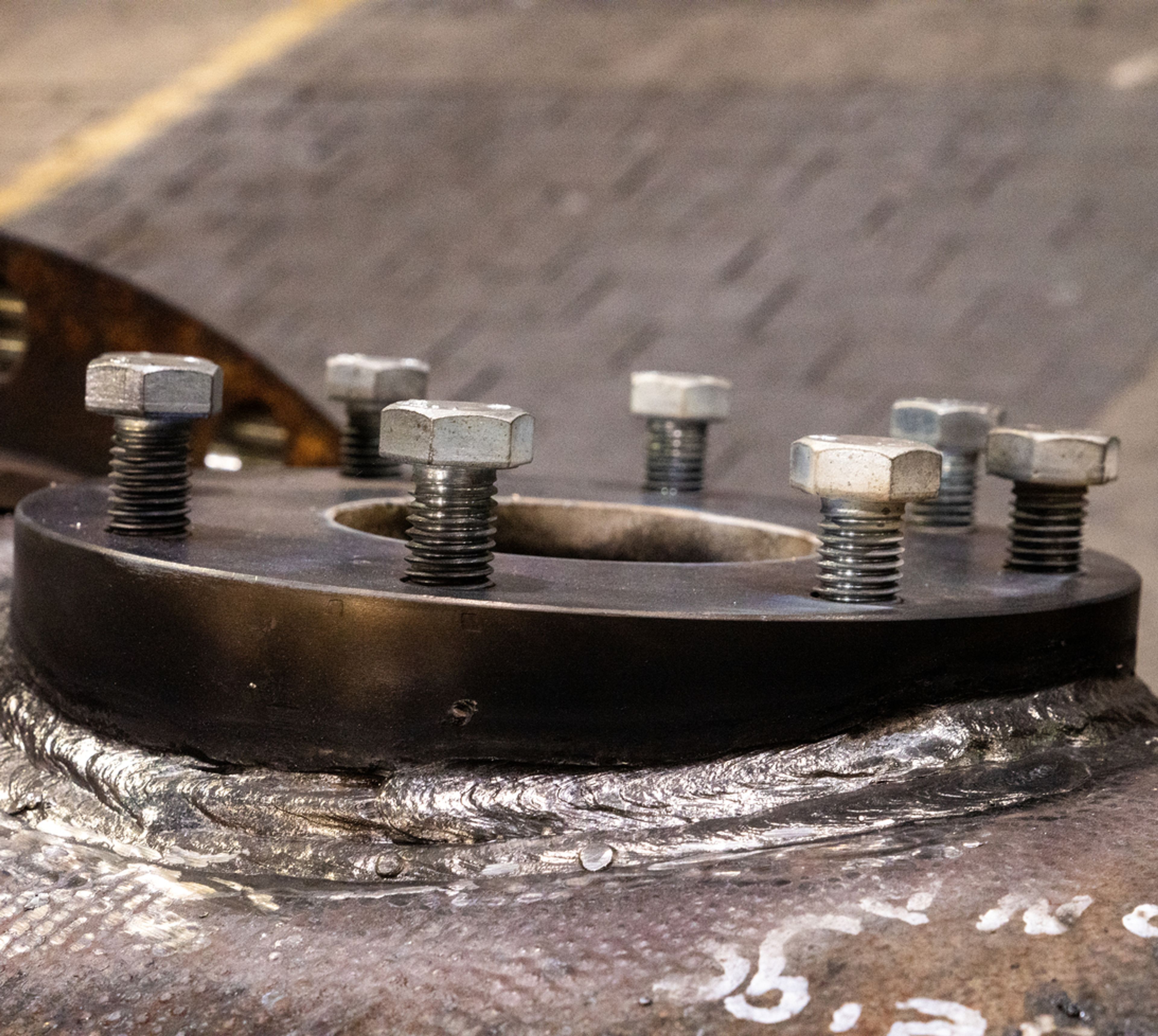 A close-up view of large metal bolts and nuts securing a circular black metal flange to a welded steel surface in an industrial setting.