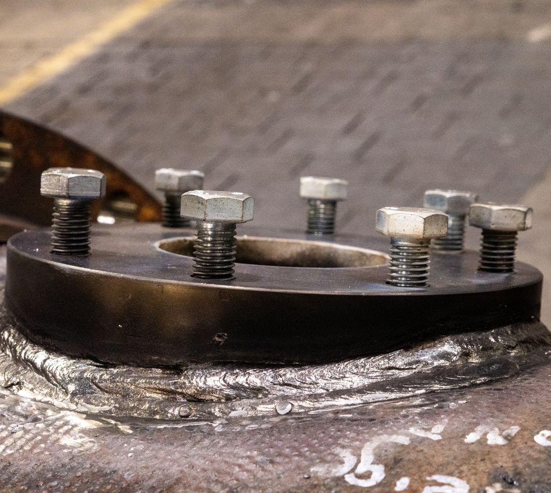 A close-up view of large metal bolts and nuts securing a circular black metal flange to a welded steel surface in an industrial setting.