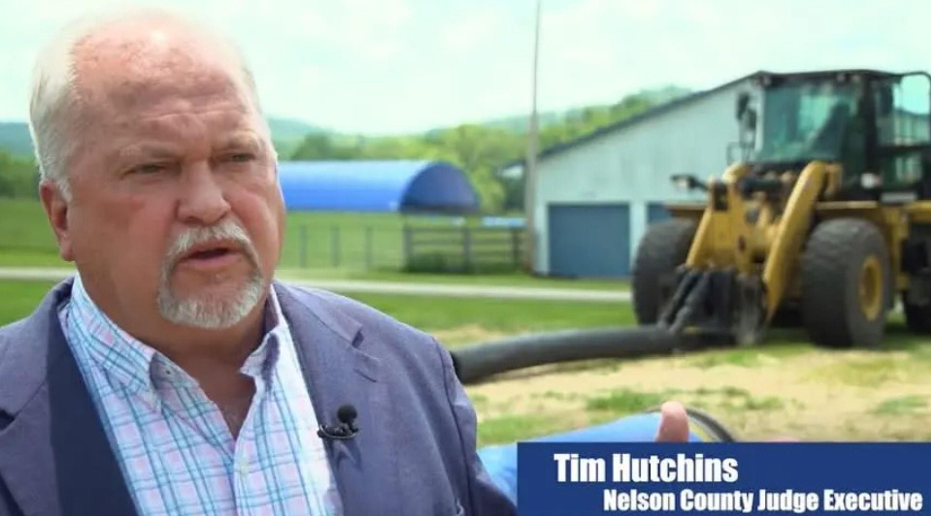 A man in a suit speaks outdoors near a construction vehicle, with buildings and fields in the background. Text on the image reads “Tim Hutchins Nelson County Judge Executive.”.