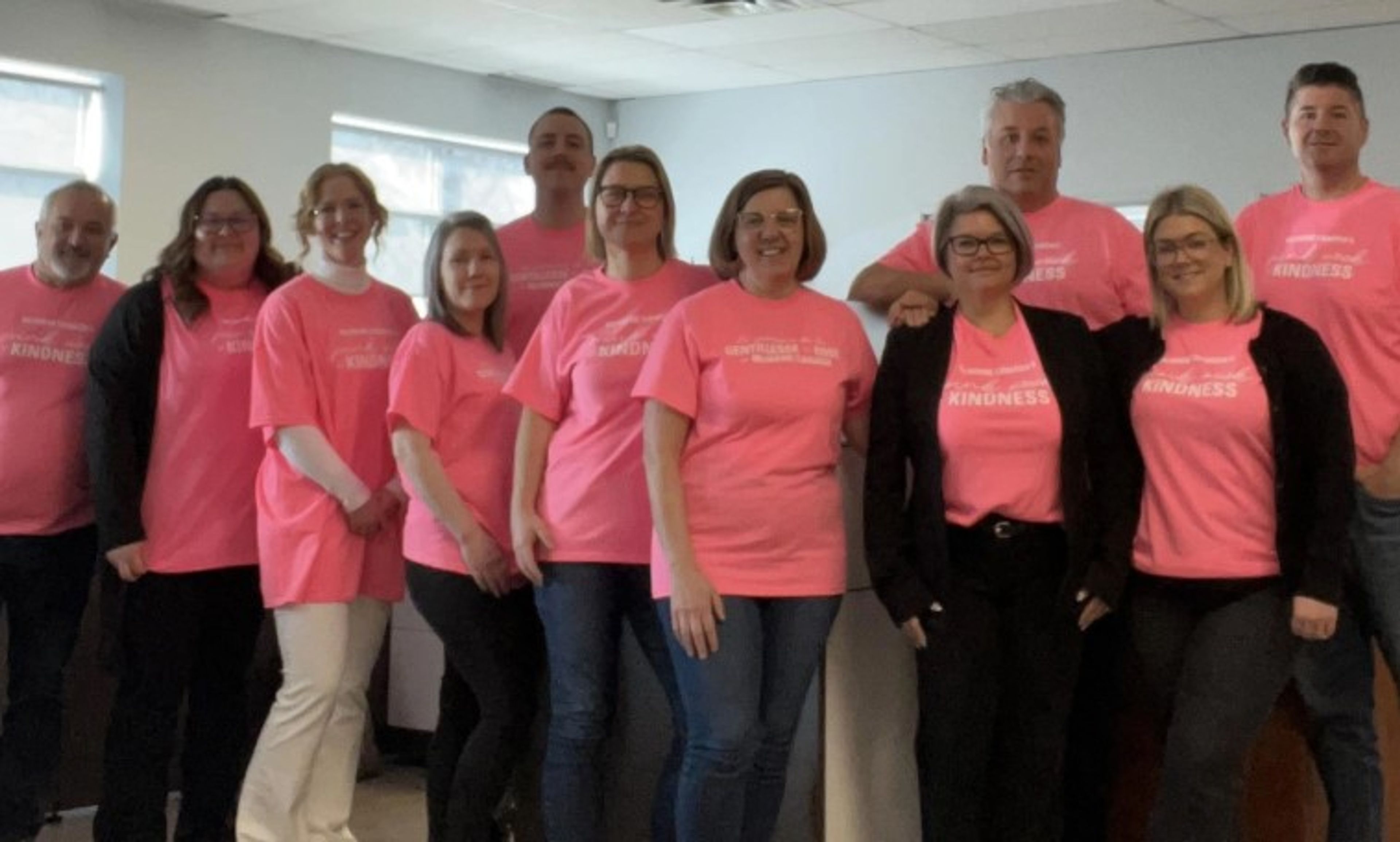 A group of eleven adults stands indoors, all wearing matching pink shirts that promote kindness. They are smiling and posing together in a well-lit room with windows in the background.