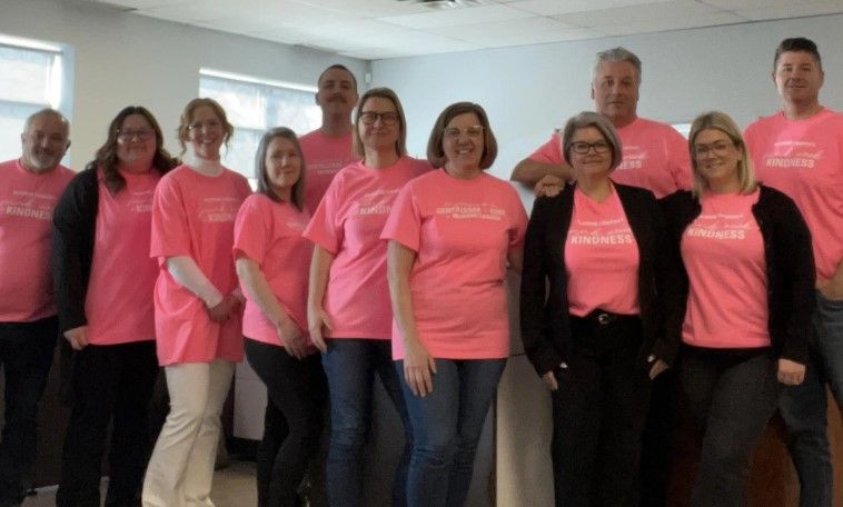 A group of eleven adults stands indoors, all wearing matching pink shirts that promote kindness. They are smiling and posing together in a well-lit room with windows in the background.