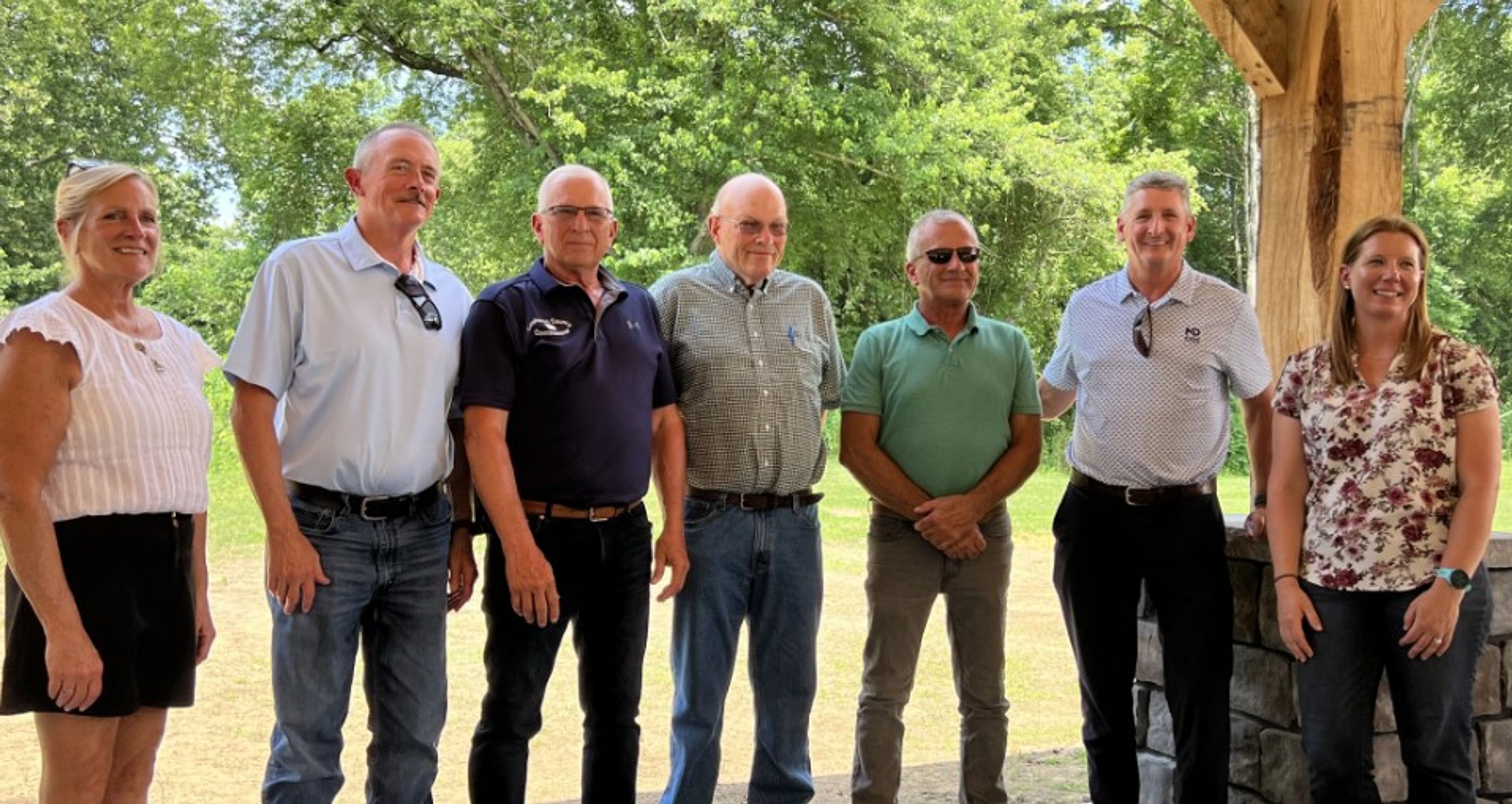 Seven adults stand side by side outdoors under a wooden structure, smiling at the camera. There are trees and greenery in the background, and the group appears to be at a casual event.