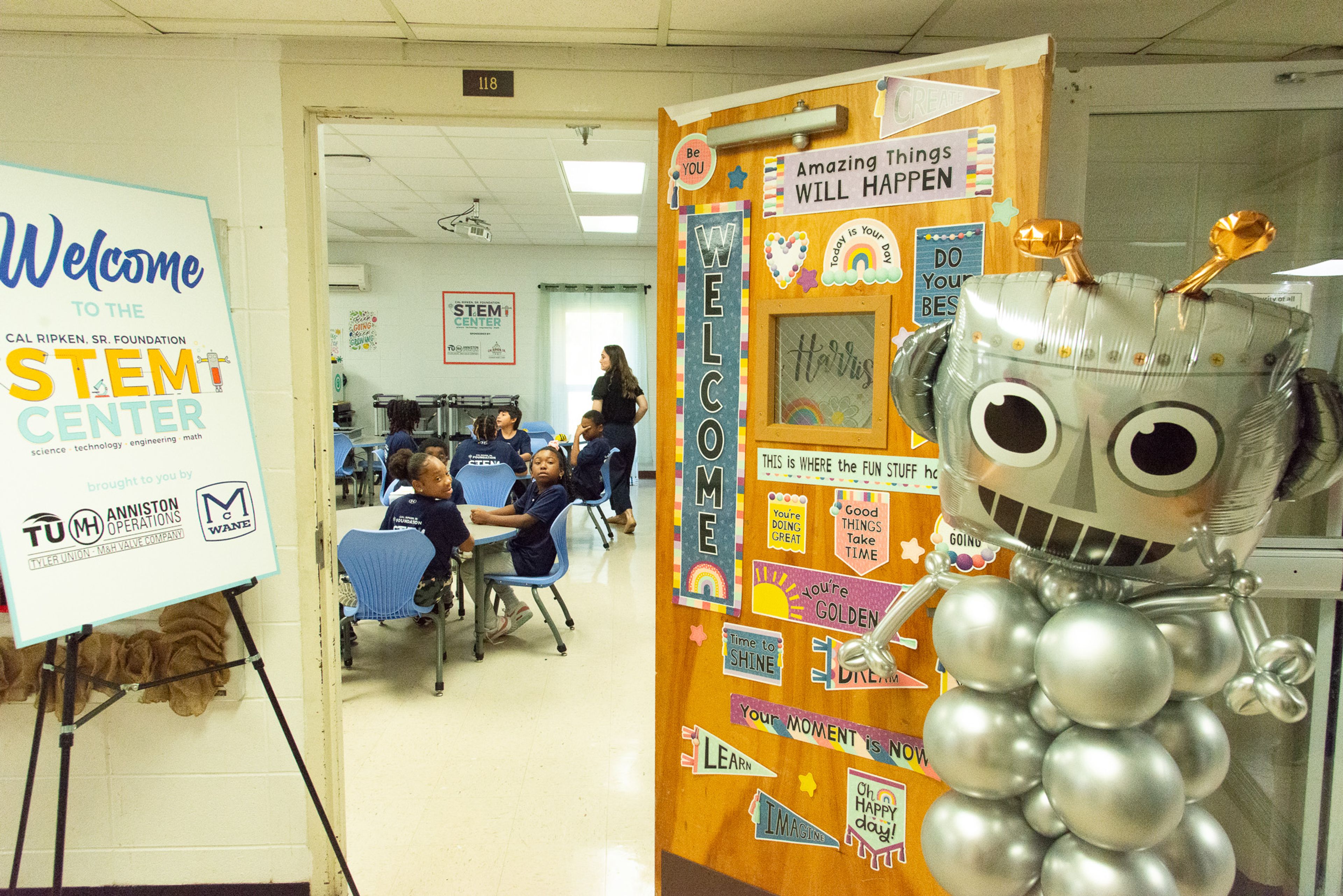 Students sit at desks in a classroom labeled “STEM Center,” with a colorful decorated door open and a silver robot balloon figure standing to the right. A welcome sign is displayed on an easel near the entrance.