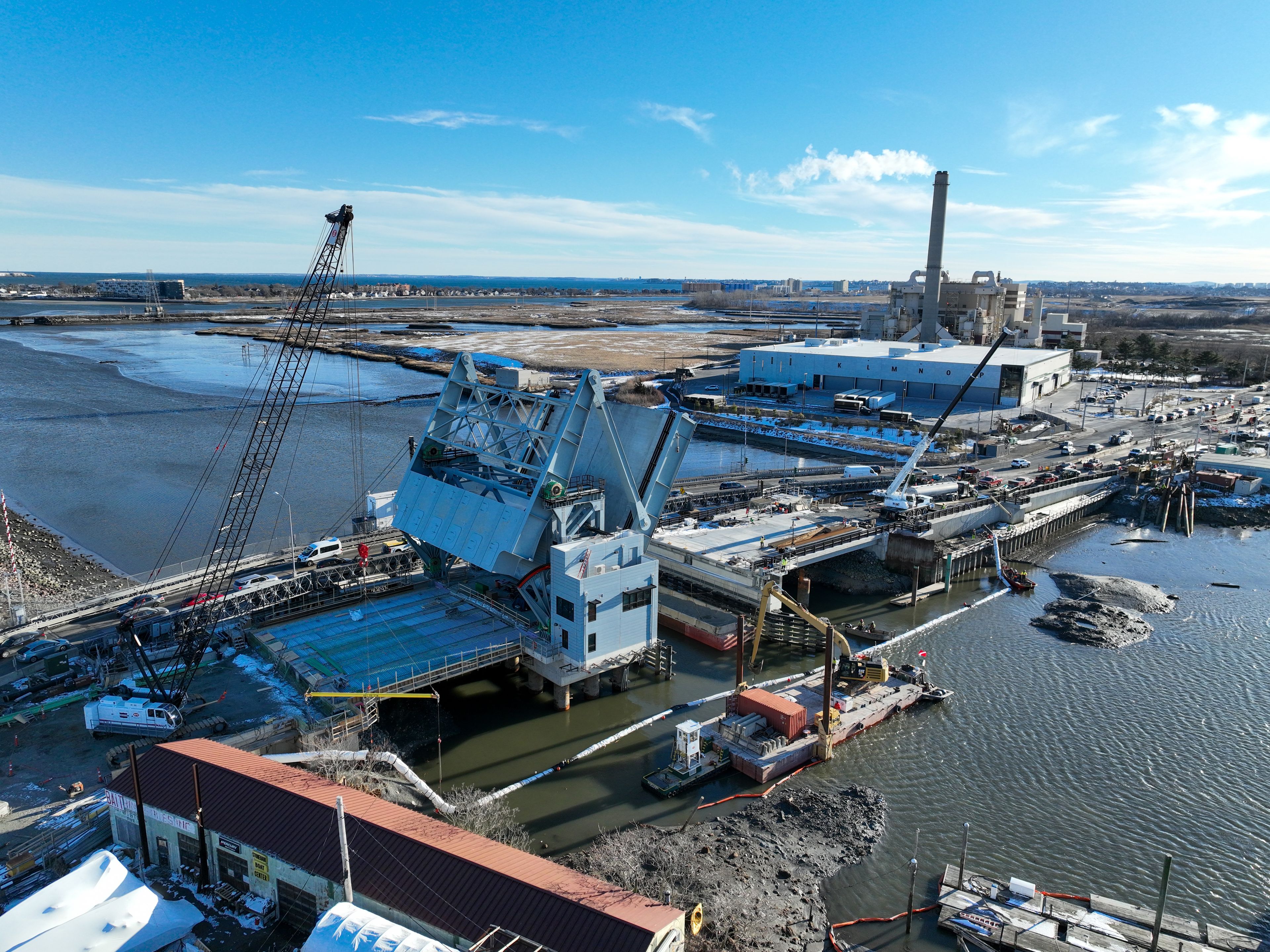 Aerial view of a drawbridge under construction over a river, with cranes, construction equipment, nearby industrial buildings, and water in the background on a clear day.