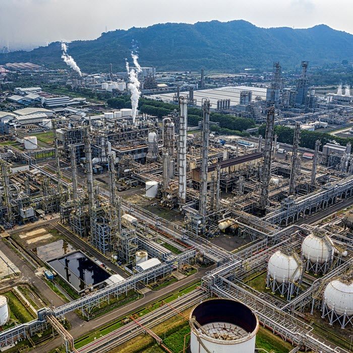 Aerial view of a large oil refinery with numerous pipes, storage tanks, and smokestacks releasing steam; green hills and industrial buildings in the background.