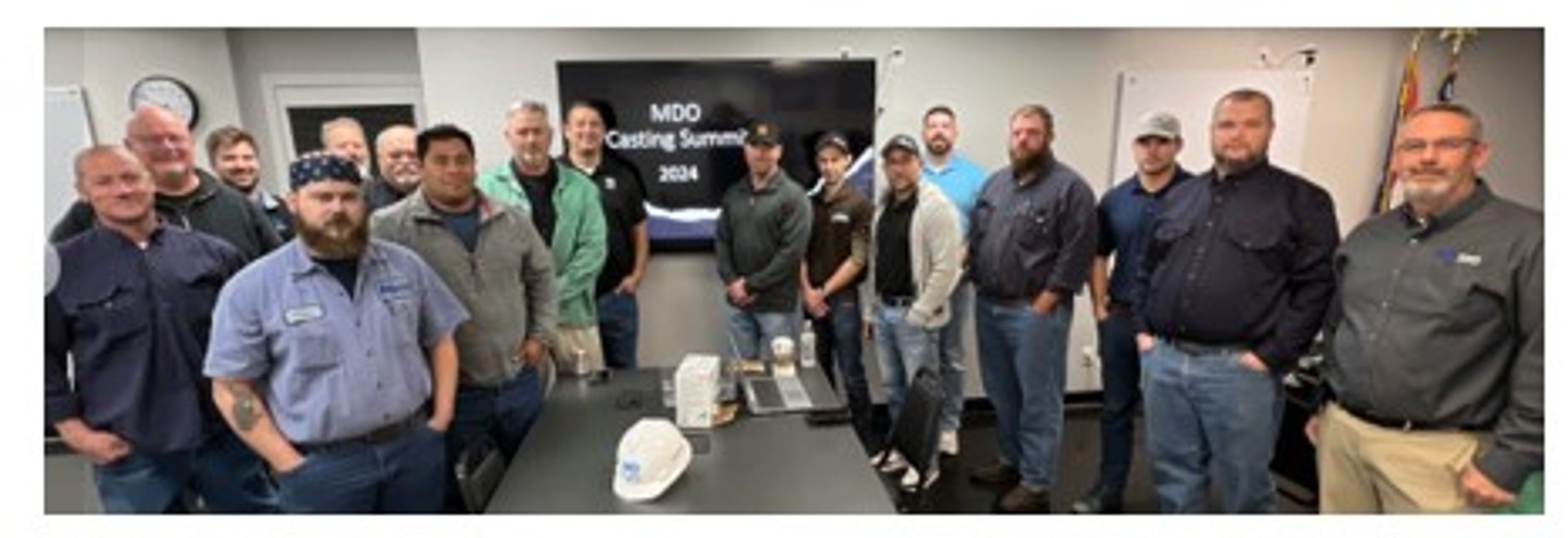 A group of sixteen men stand together in a meeting room with a screen reading “MDO Coating Summit 2024” in the background. There are coffee cups and a hard hat on the table in front of them.