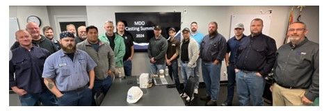 A group of sixteen men stand together in a meeting room with a screen reading “MDO Coating Summit 2024” in the background. There are coffee cups and a hard hat on the table in front of them.
