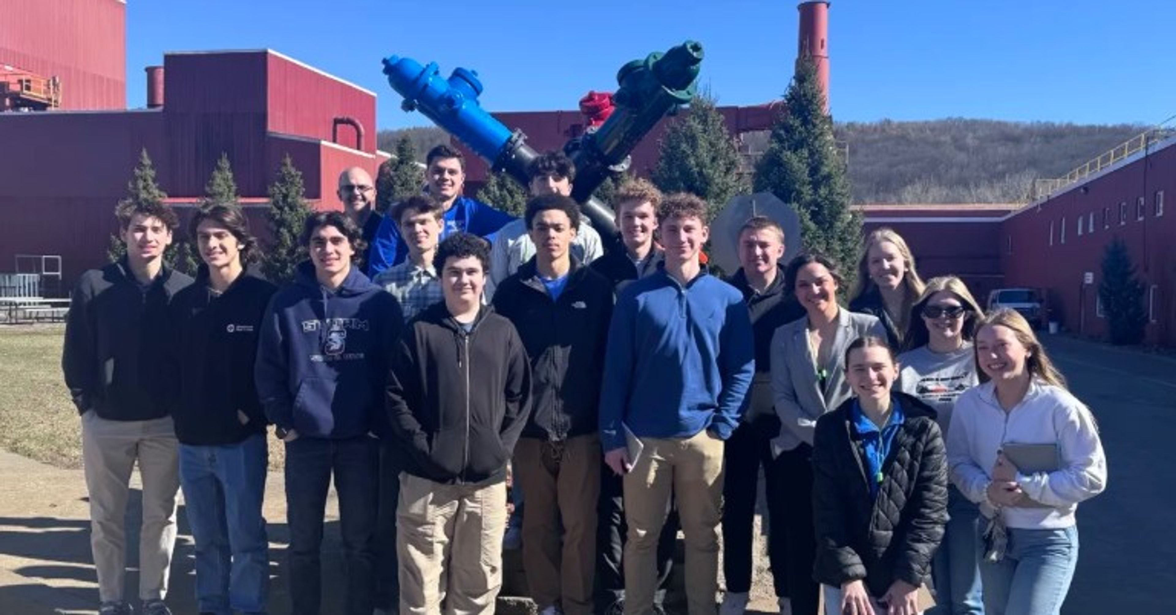 A group of sixteen people stands outside in front of red industrial buildings and evergreen trees on a sunny day. Some are smiling, and all are dressed in casual, cool-weather clothing.
