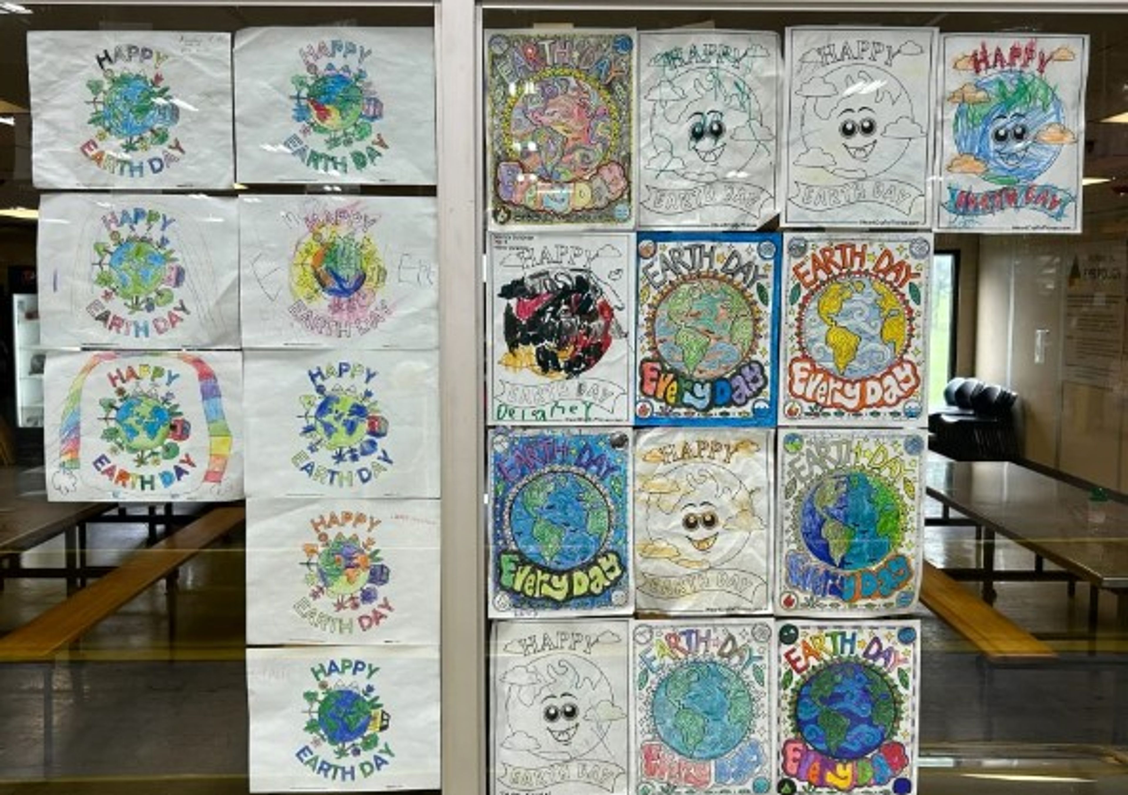Colorful Earth Day drawings and posters, made by children, are displayed on glass. Each artwork features the Earth with a smiley face, “Happy Earth Day” messages, and various creative designs and colors.